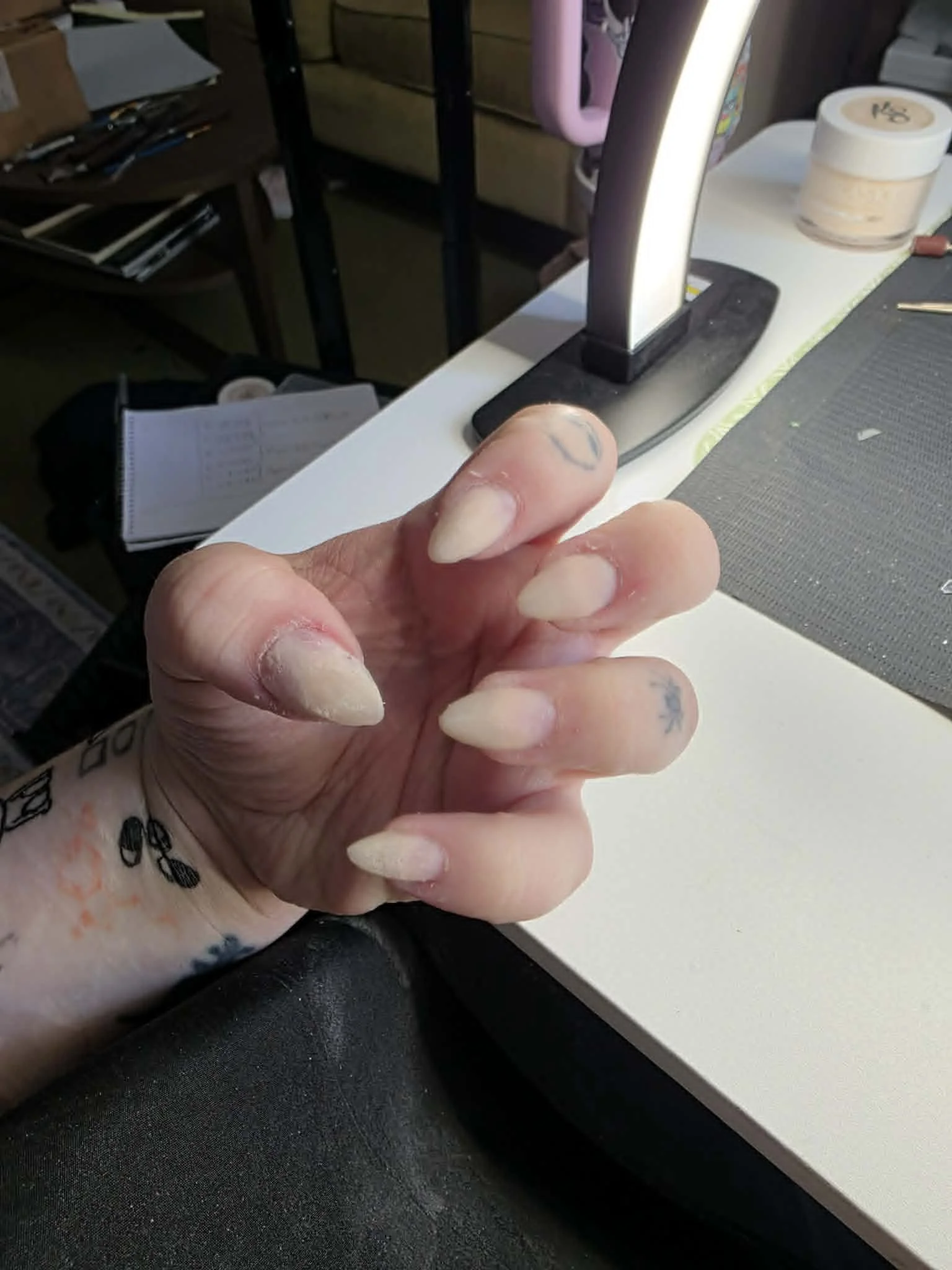 Close-up of a person's hand with long, pointed fingernails, slightly dirty, with tattoos on fingers and wrist, pointing near a desk lamp on a white desk surface. The background includes various items such as a notebook, a laptop, and containers.