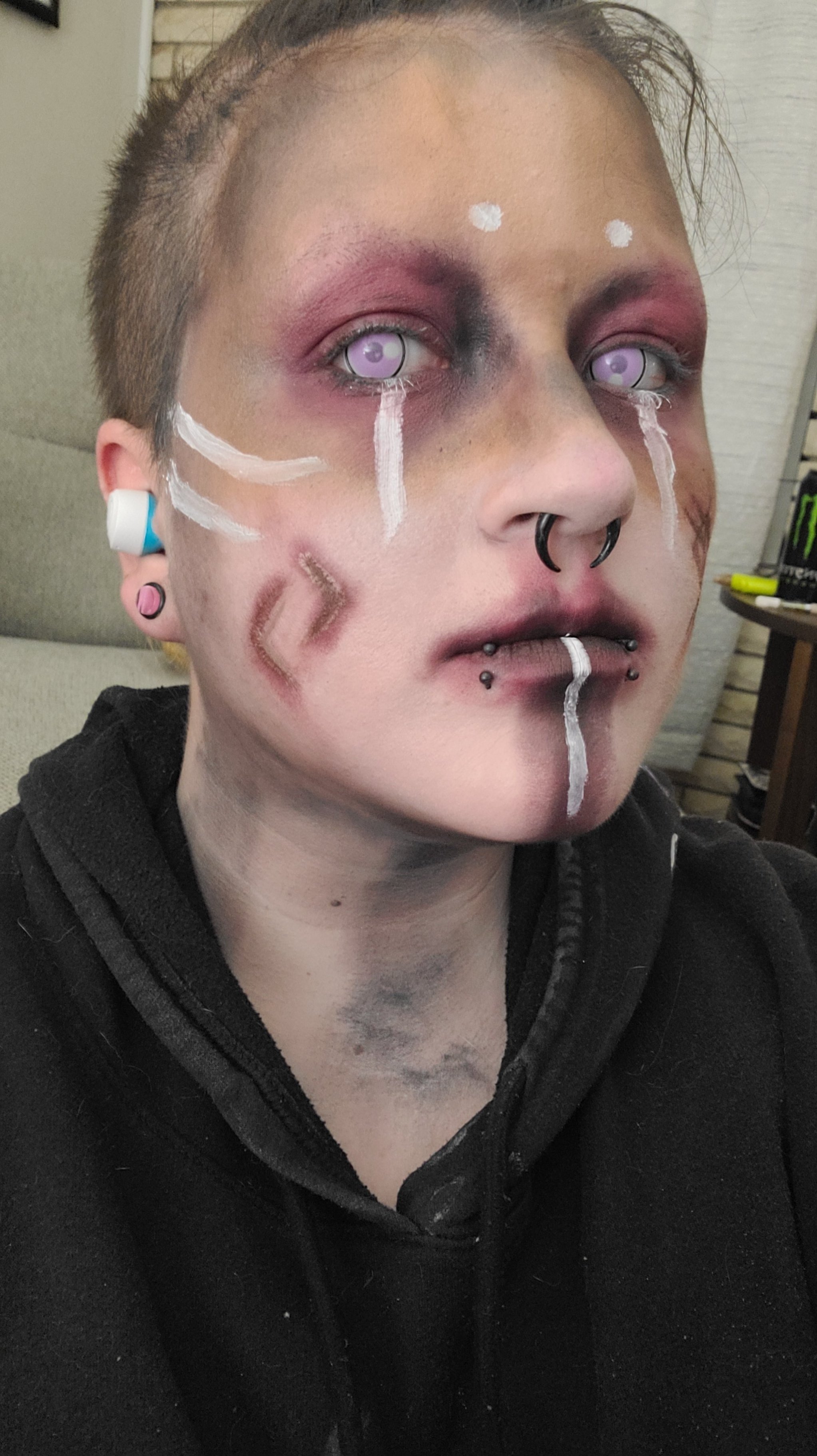A person with short hair, wearing makeup with purple contact lenses, black and purple face tattoos, and multiple piercings, including septum and lip piercings. They are dressed in a black hoodie and sitting indoors.