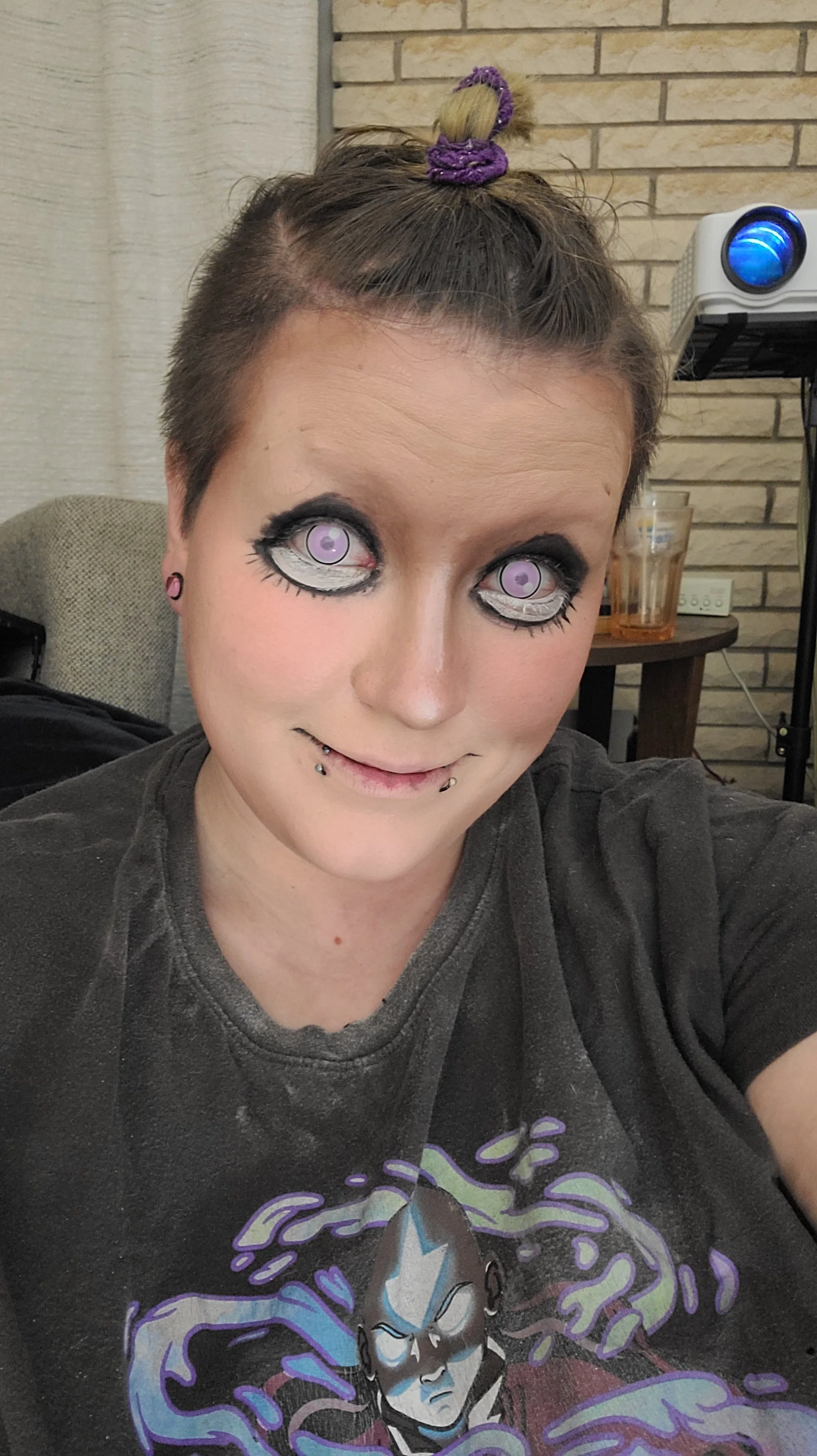 A person with short brown hair styled in a top knot with a purple hair scrunchie, wearing dramatic eye makeup with lavendar contact lenses, multiple facial piercings, and a graphic t-shirt featuring a stylized superhero character with a purple and bl