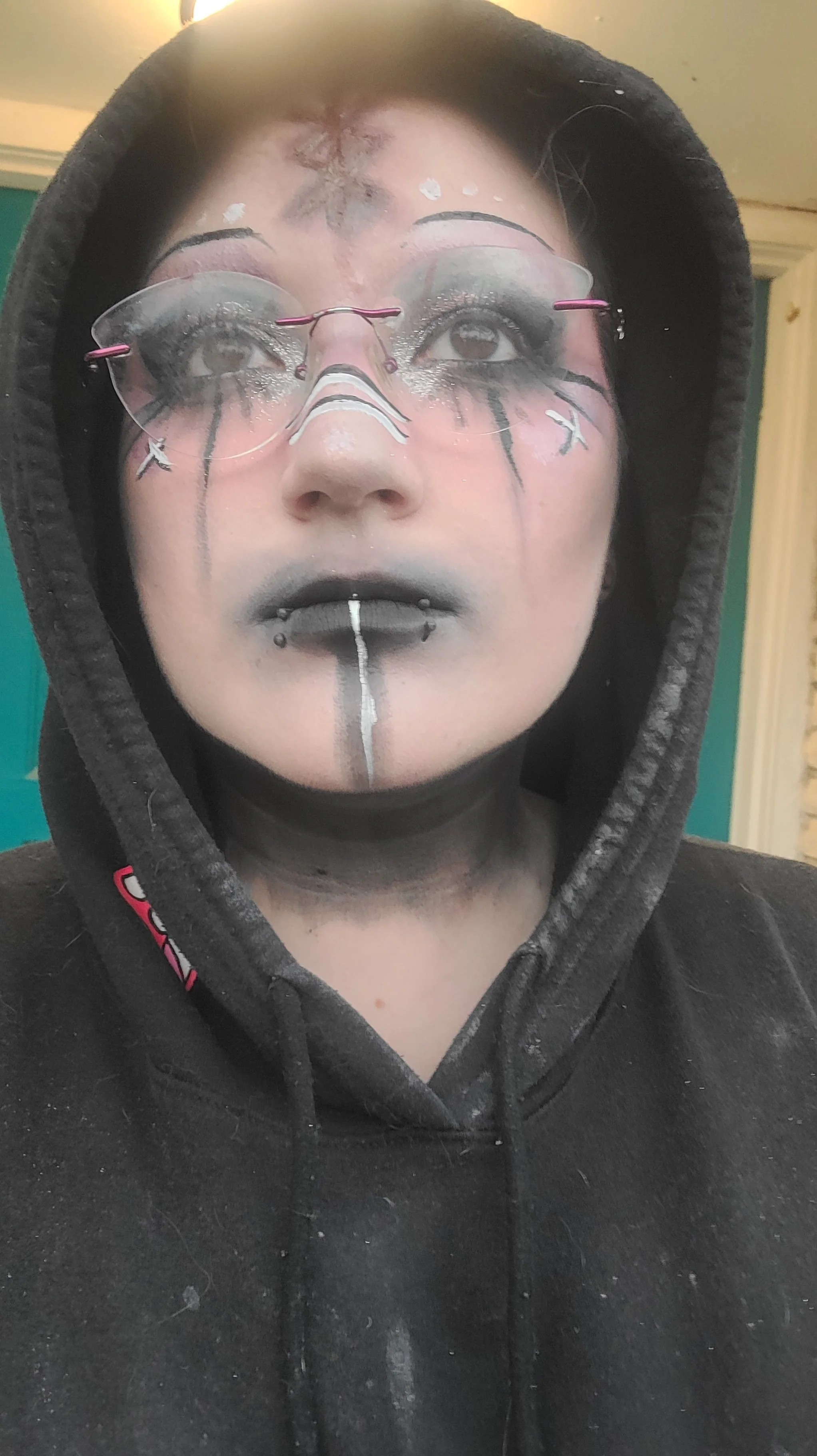 Person wearing a black hoodie and glasses with makeup resembling a dragon, featuring black, white, and silver details, including a line running down the chin and black lipstick.