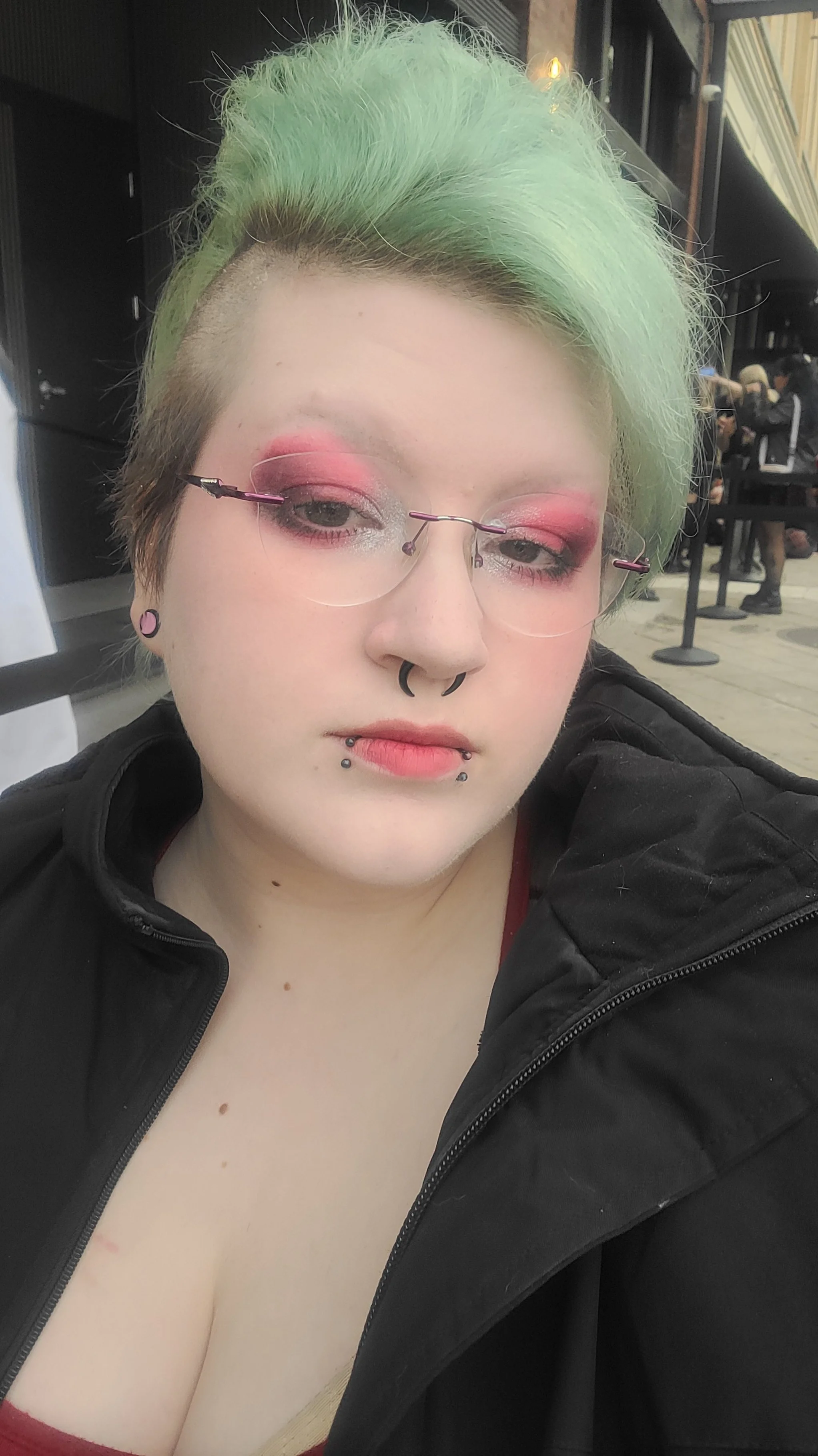 A woman with short, green hair and glasses, wearing pink eyeshadow and a black jacket, taking a selfie indoors.