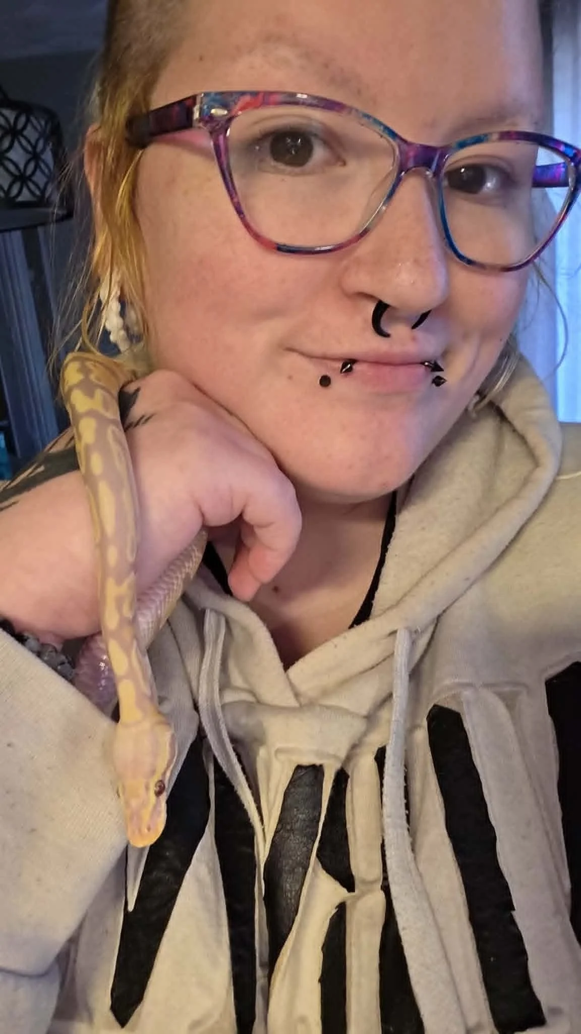 A person with blond hair and glasses holding a small snake in their hand, wearing a beige hoodie with black stripes.