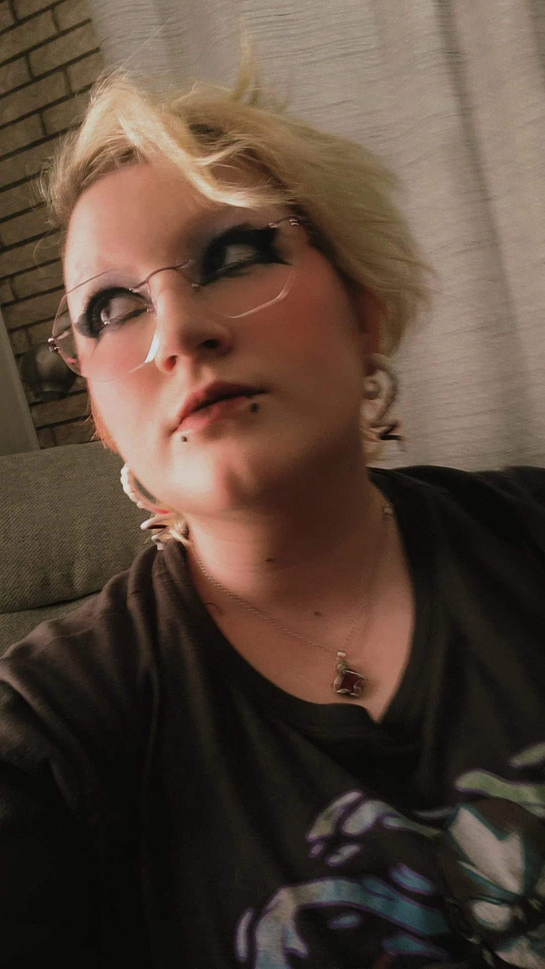 A young person with short blonde hair, wearing glasses, a black graphic T-shirt, and jewelry, sitting on a couch with a brick wall in the background.