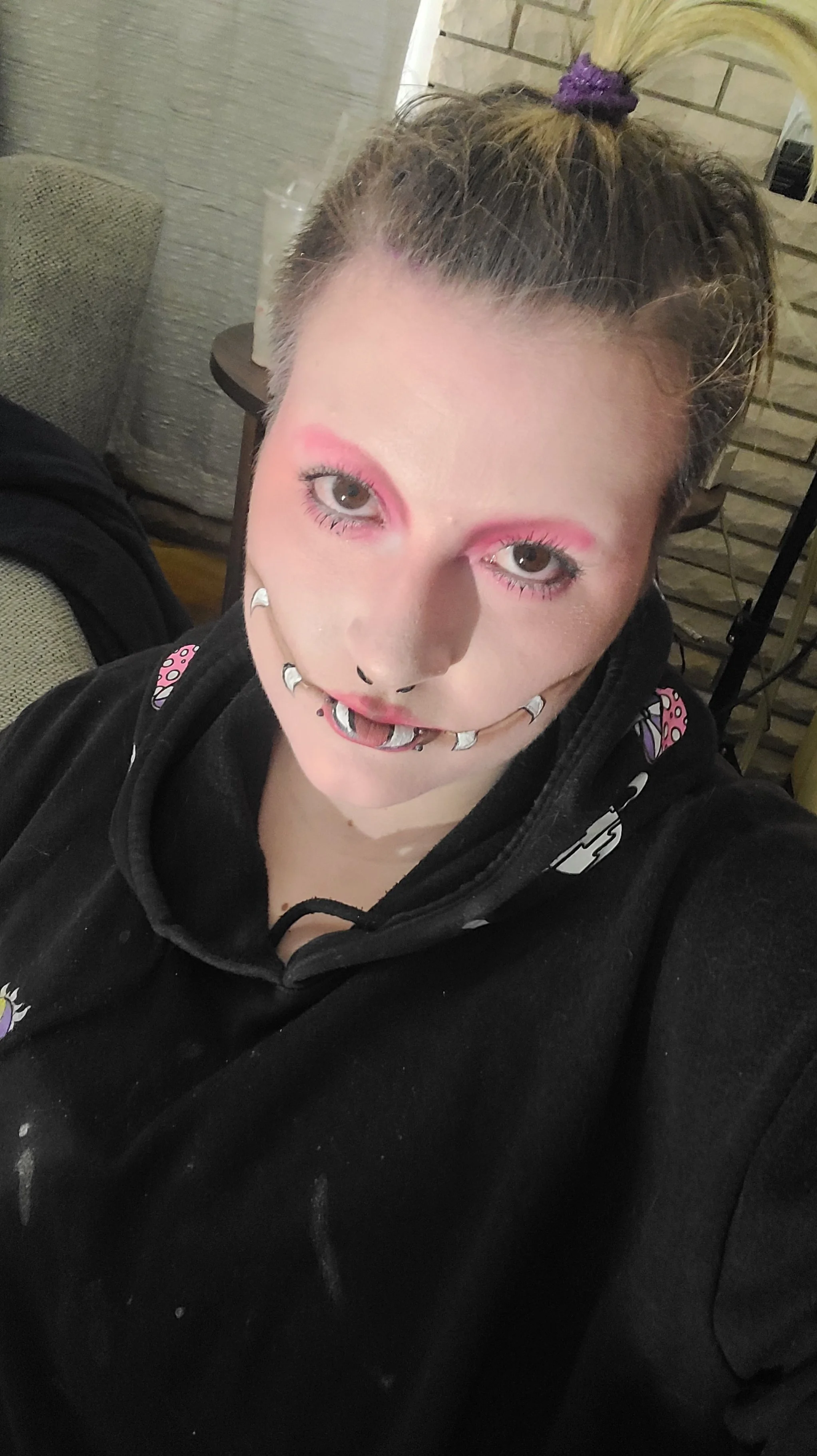 A young woman with light skin, brown eyes, and freckles, wearing makeup with pink eyeshadow, and sporting multiple facial piercings, including a septum ring, lip piercings, and earrings. Her hair is pulled up into a tied bun with a purple scrunchie. 