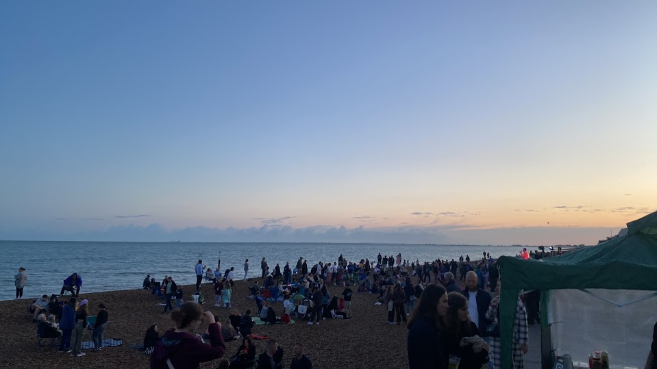 People gathered on a sandy beach during sunset, some sitting and others walking, with a green tent on the right side and the ocean in the background.