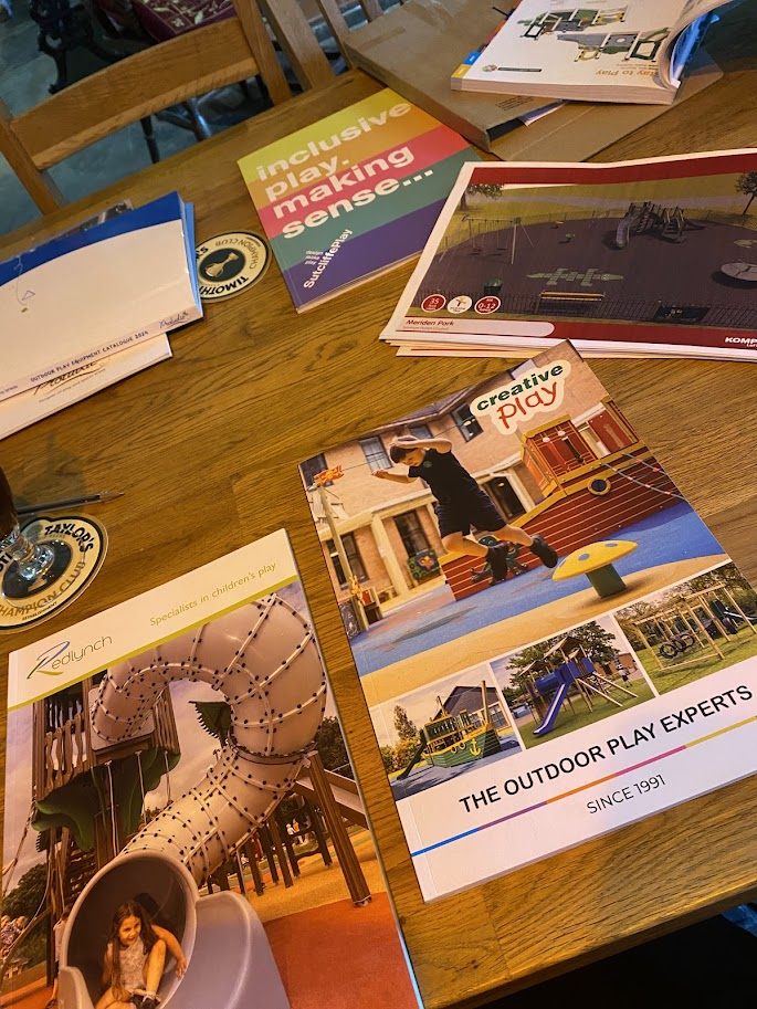 A wooden table with various brochures and magazines about children's play and outdoor playgrounds. One brochure features a girl climbing on playground equipment, another shows a slide, and others have colorful covers and images related to outdoor play spaces.