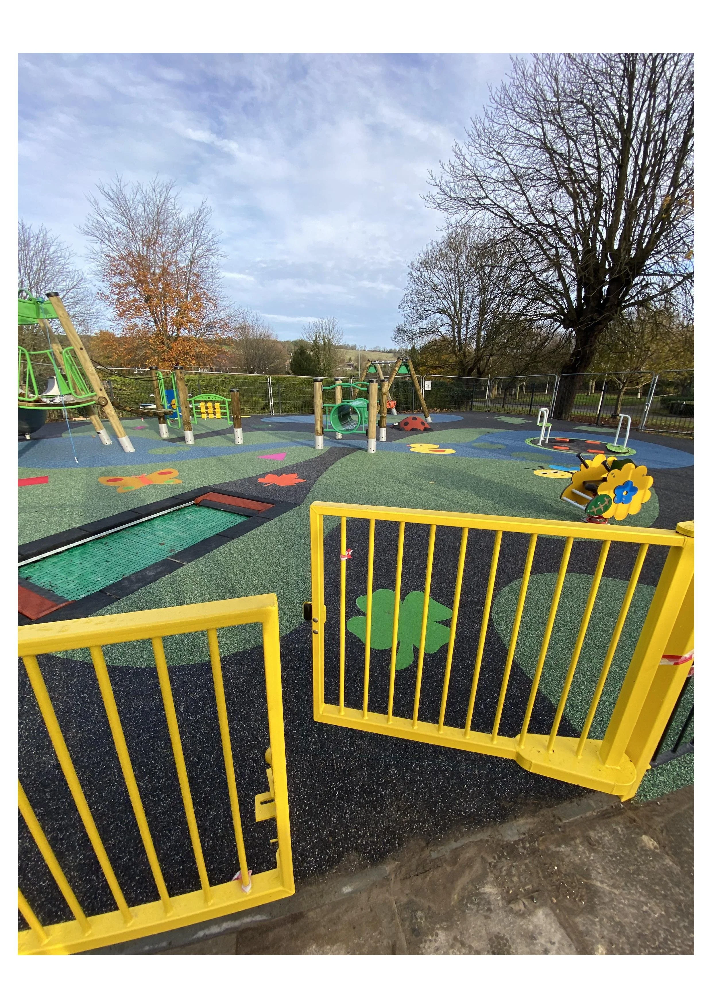 Colorful playground with play structures, including slides, a climbing frame, and spring riders, enclosed by yellow metal gates, on a rubber surface with decorative flower and leaf patterns. Trees with bare branches are visible in the background under a partly cloudy sky.