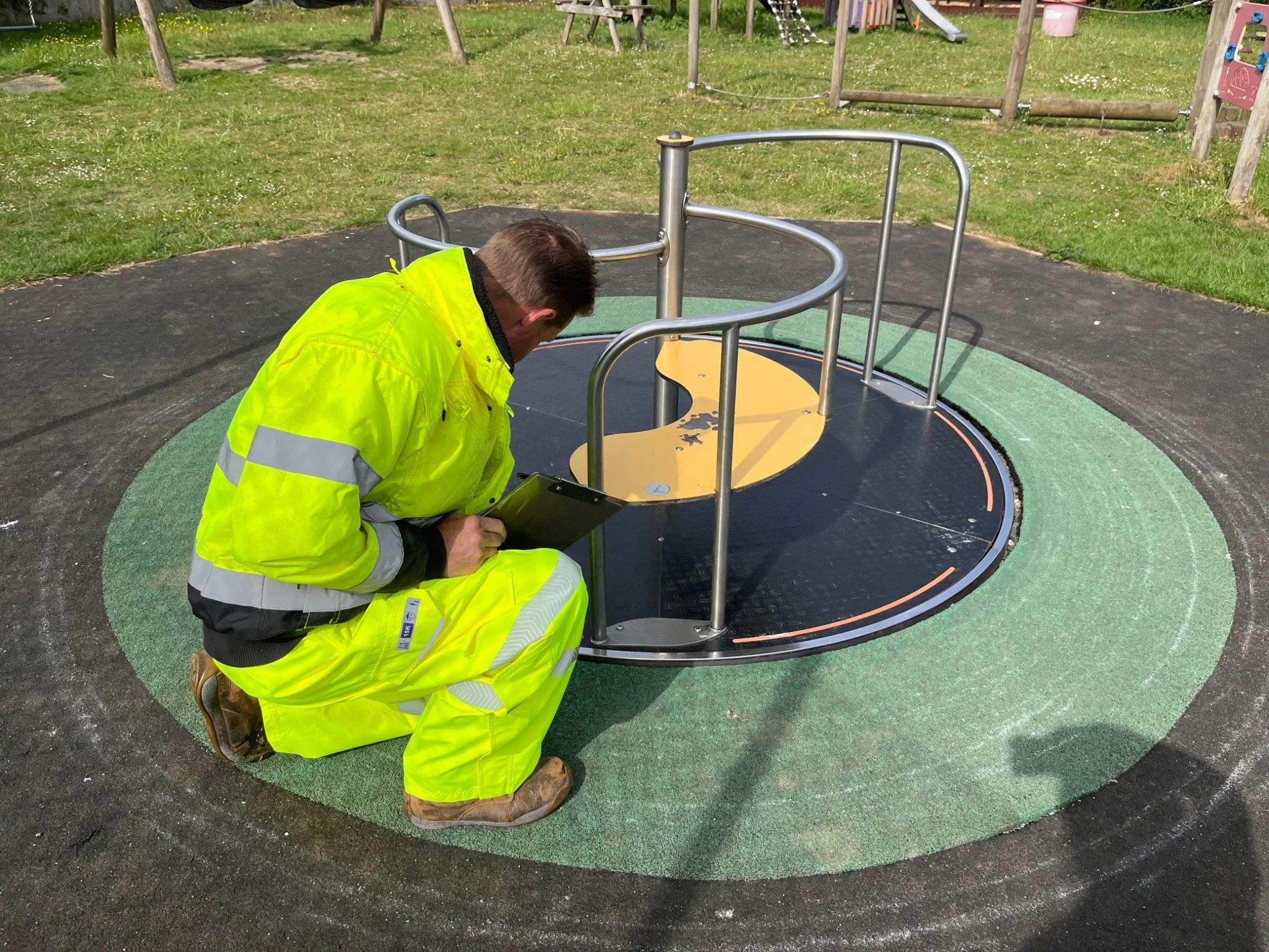 A worker wearing a high-visibility yellow jacket and pants kneeling on a playground rubber surface while inspecting or repairing a merry-go-round.