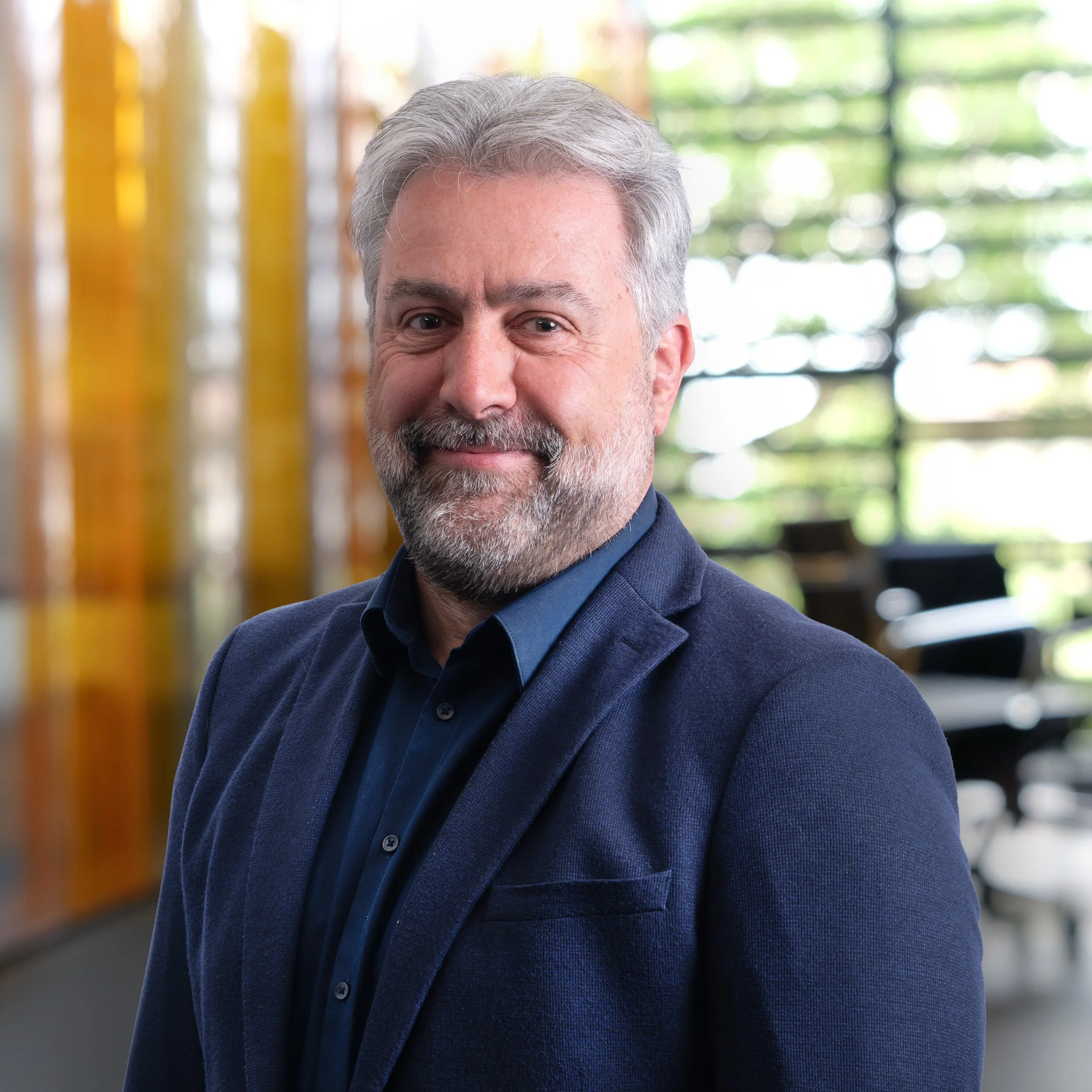 Close-up of a middle-aged man with gray hair and beard, wearing a dark blue blazer and shirt, standing outdoors with modern architectural elements in the background.