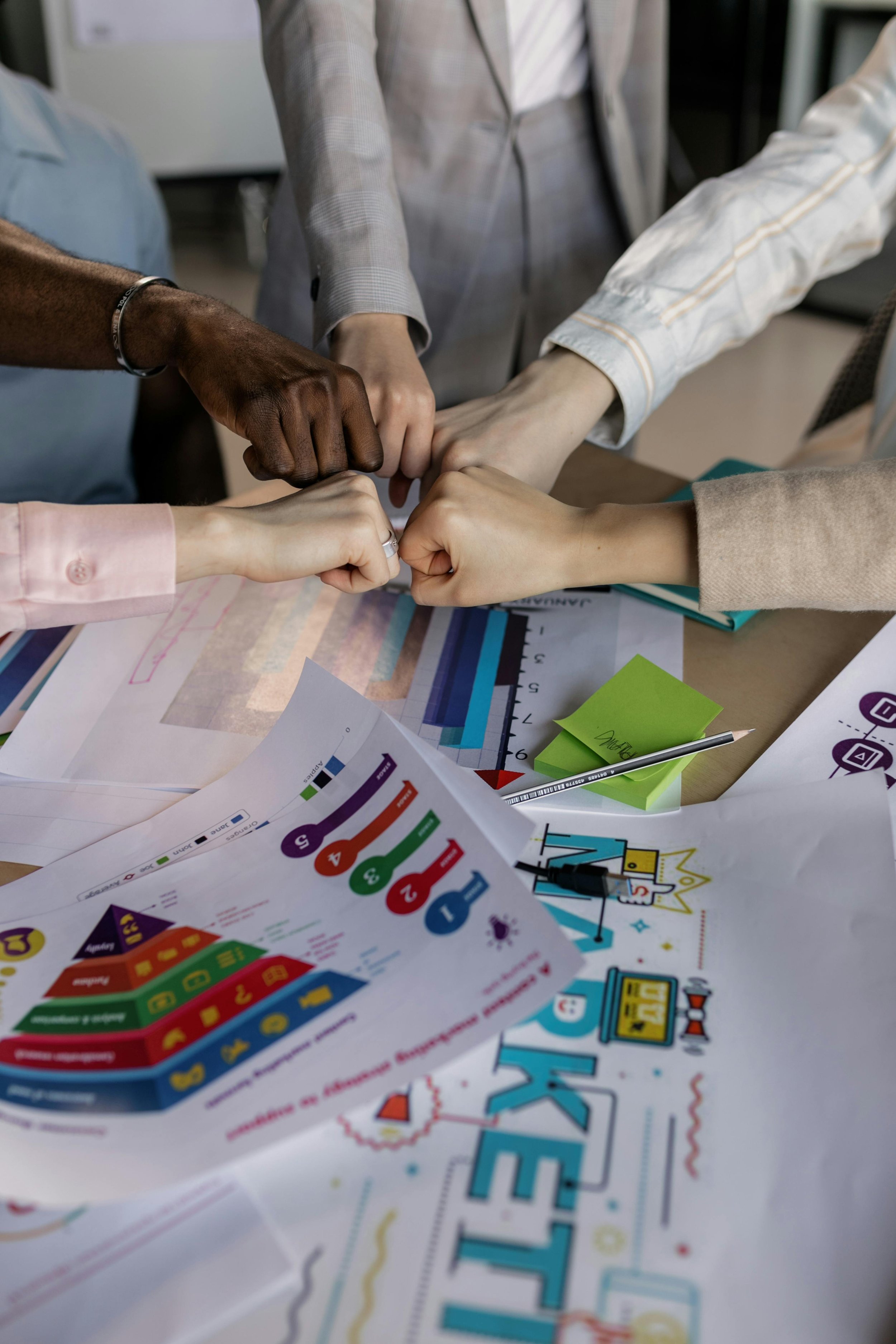 Multiple people placing fists together above a table with colorful papers, charts, and a pen.
