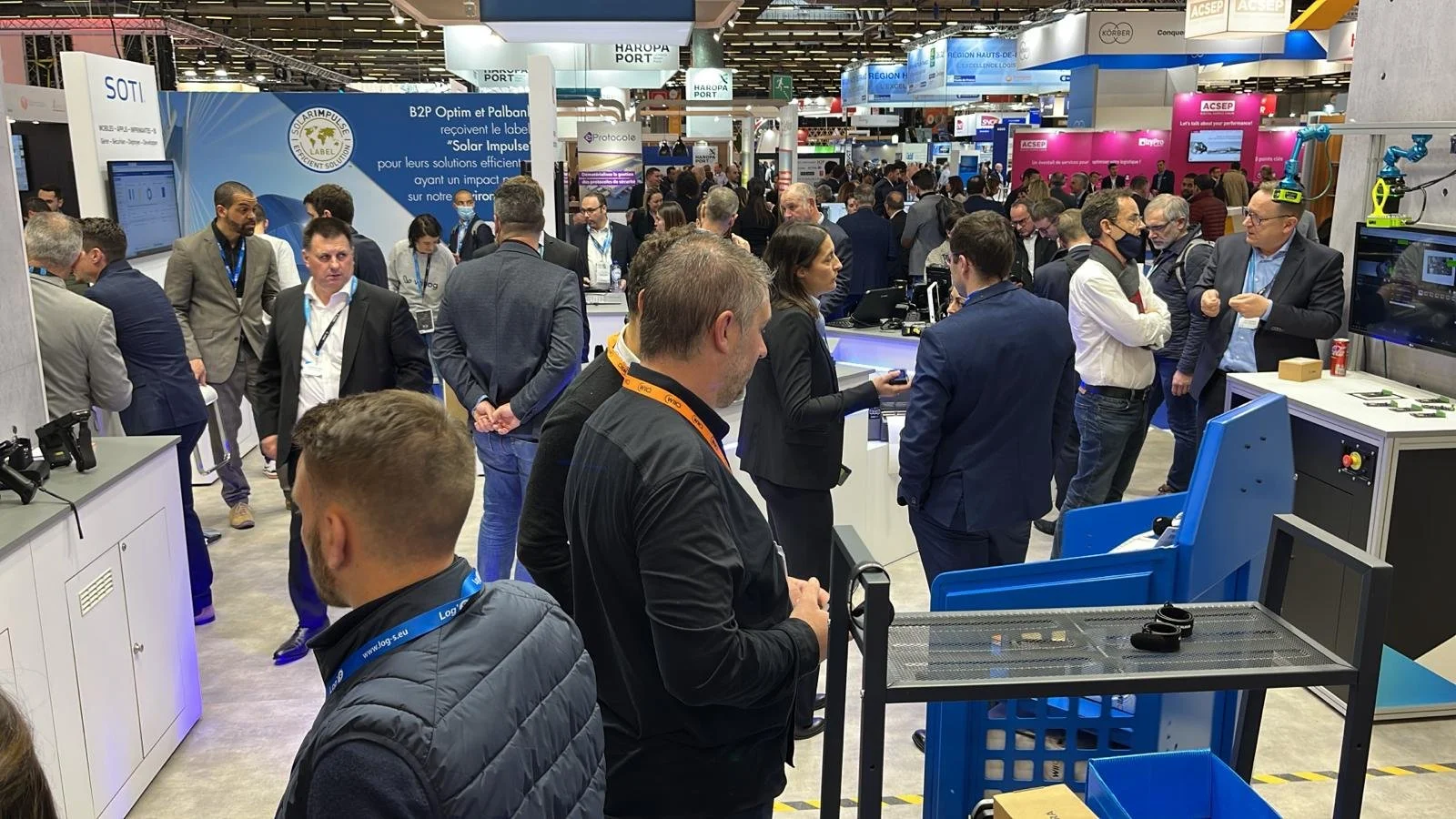 A crowded trade show or conference with many people engaging around various booths and displays. Some individuals are wearing business attire, and the background features banners and signs for different companies, with tech and industrial equipment visible.
