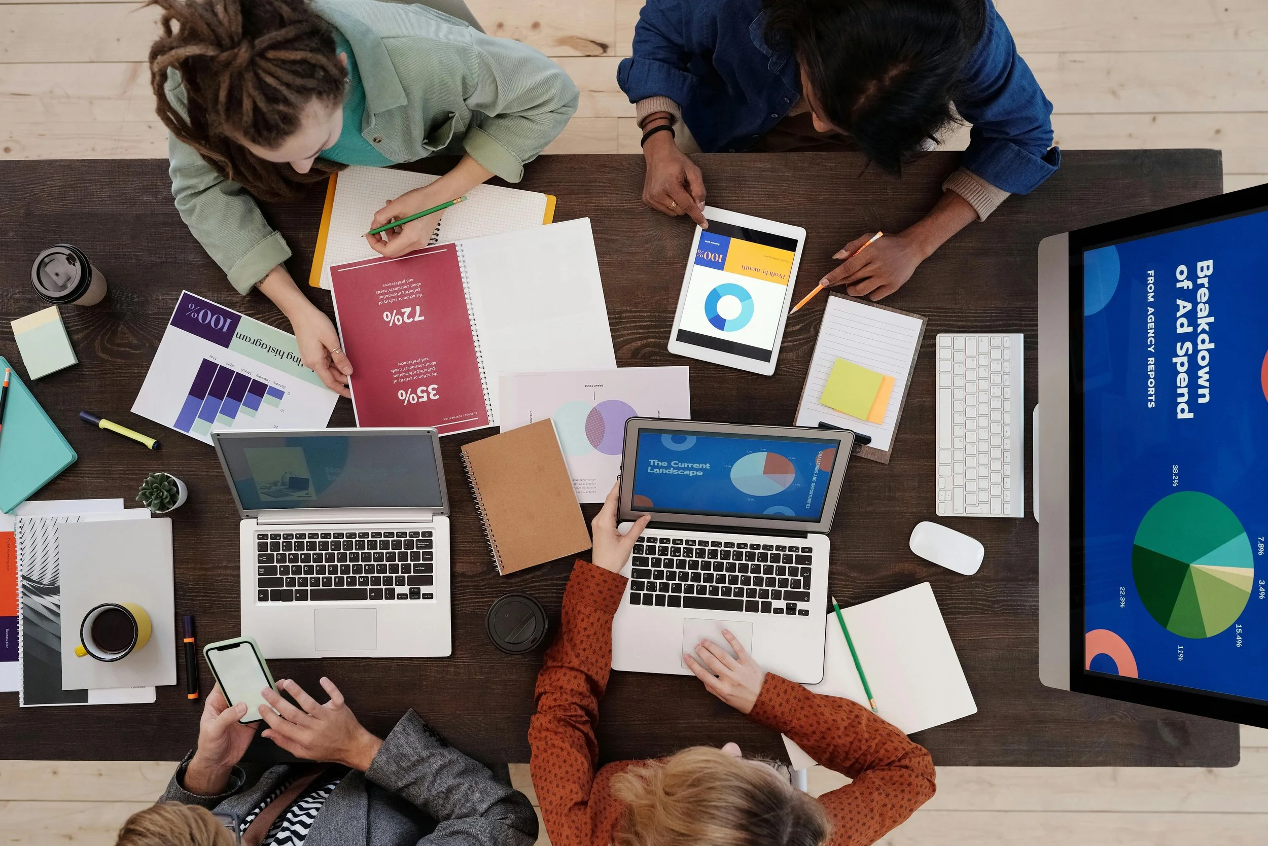 Top-down view of a group of five people gathered around a table working on laptops, tablets, and phones with various charts and graphs displayed on screens and papers.