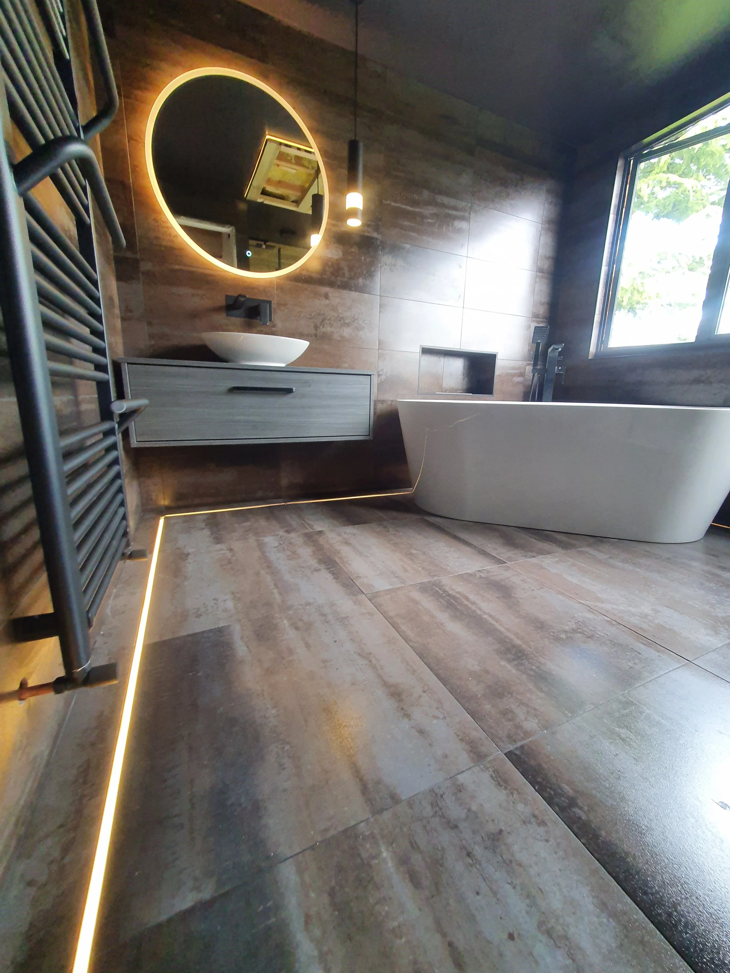 Modern bathroom with a freestanding bathtub, a wall-mounted vanity with a vessel sink, a circular backlit mirror, a window showing greenery outside, and wood-look tiled walls and floors.
