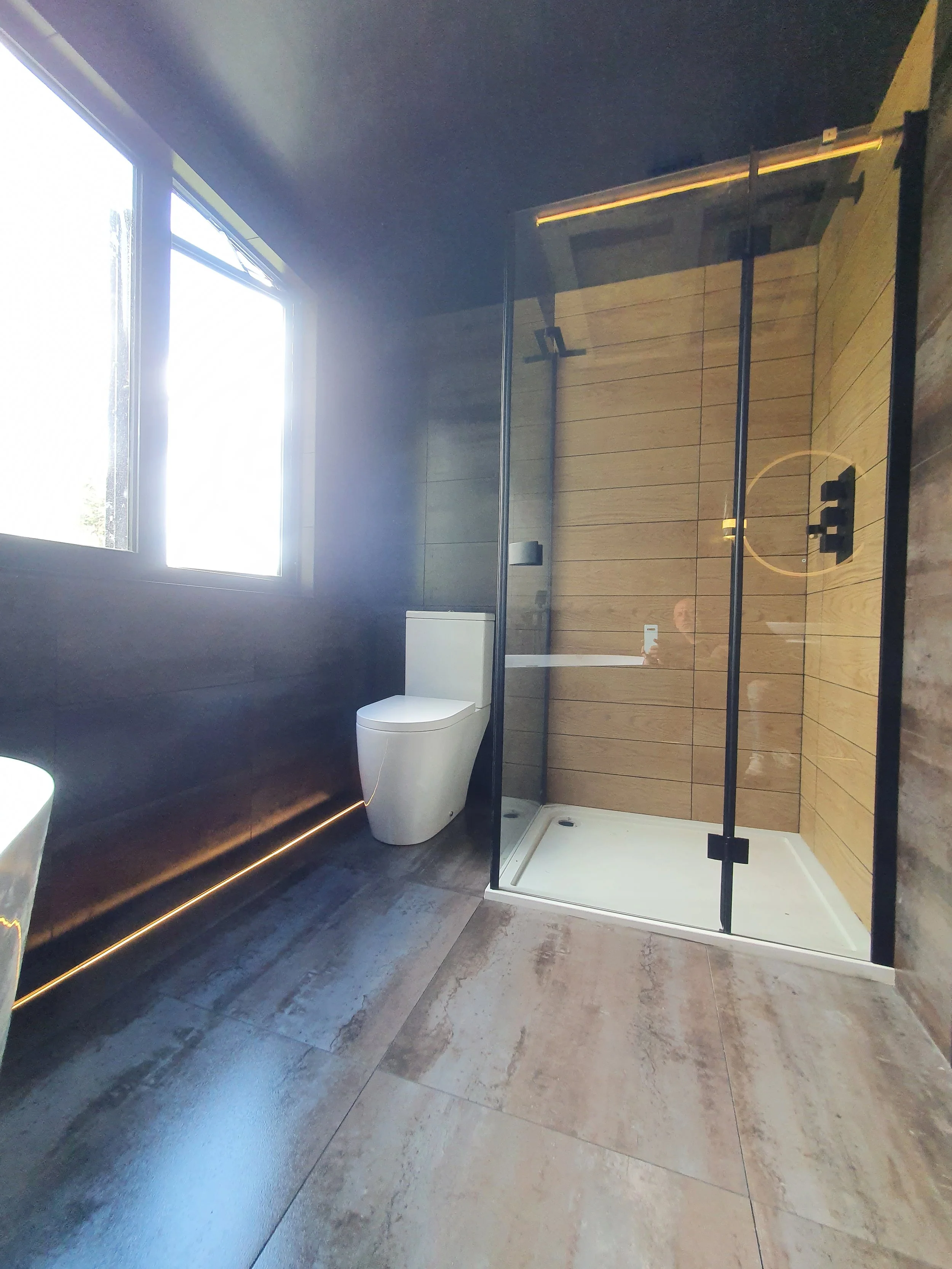 Modern bathroom with wooden tile walls, a white toilet, a glass shower enclosure with black framing, and a window allowing natural light.