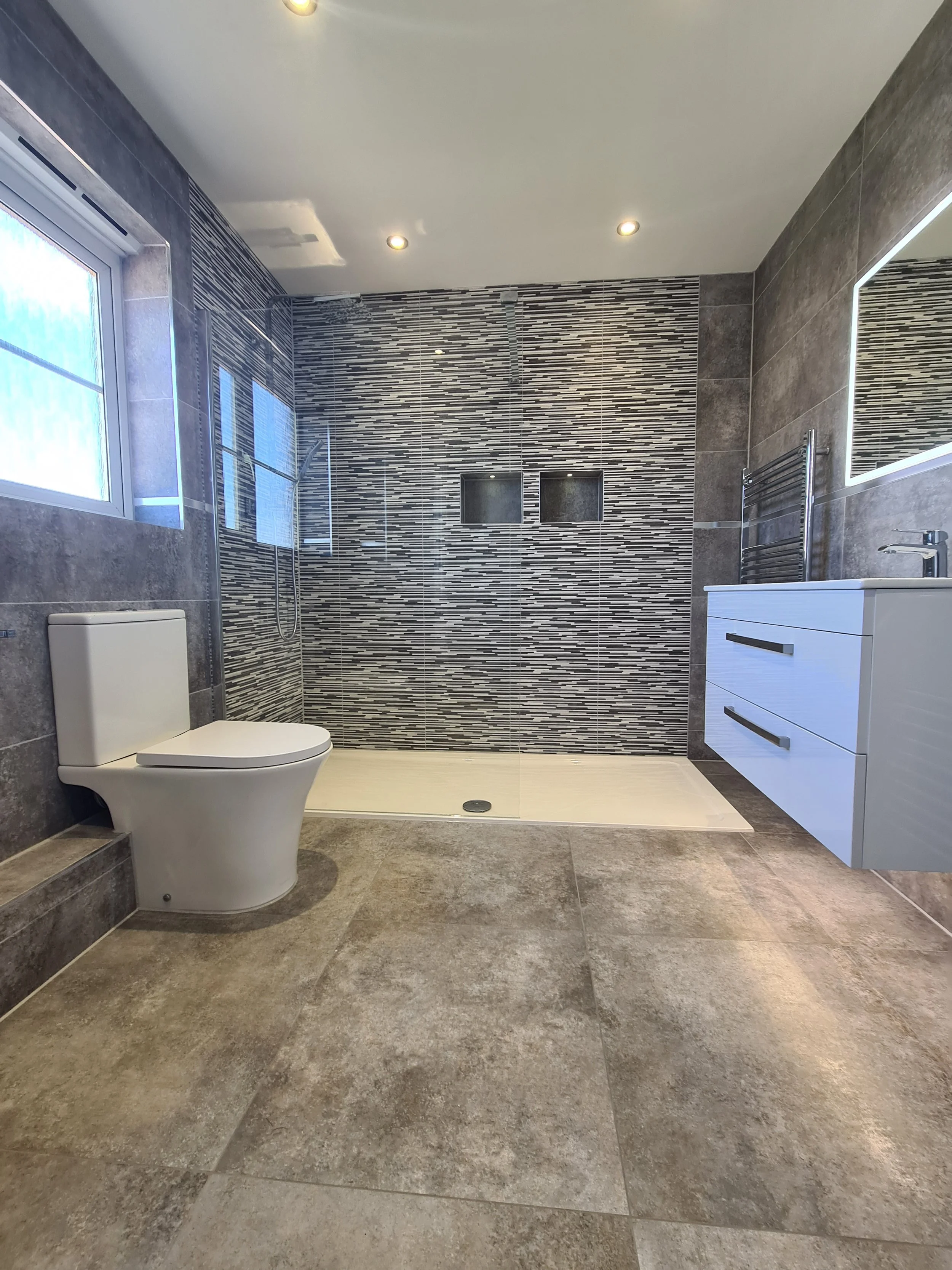 A modern bathroom with a toilet, a vanity with a mirror, and a walk-in shower with tiled walls, a window, and built-in shelves in the shower area.