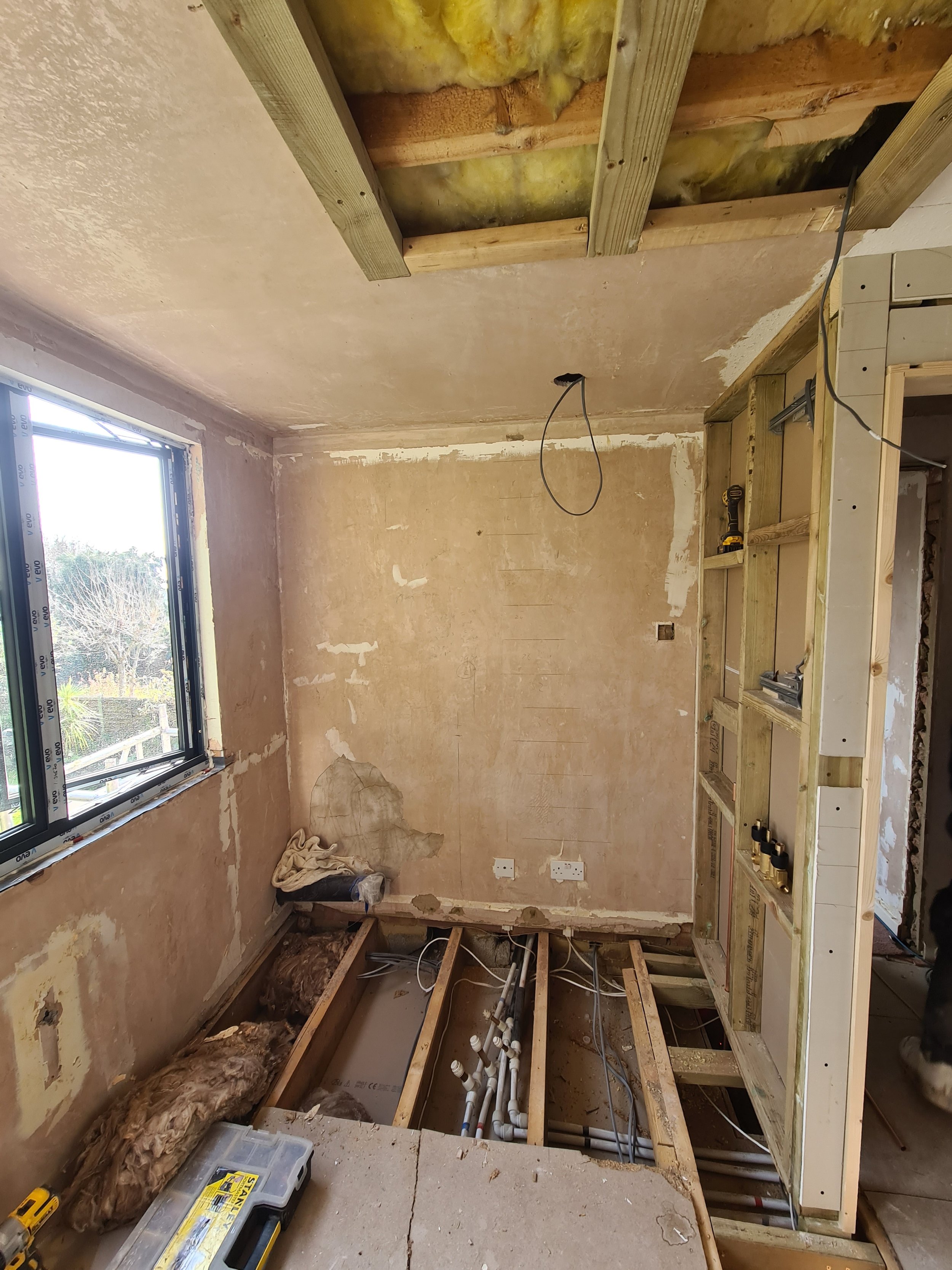 A room under renovation with exposed drywall, plumbing pipes, framing, and insulation, with a window letting in natural light.