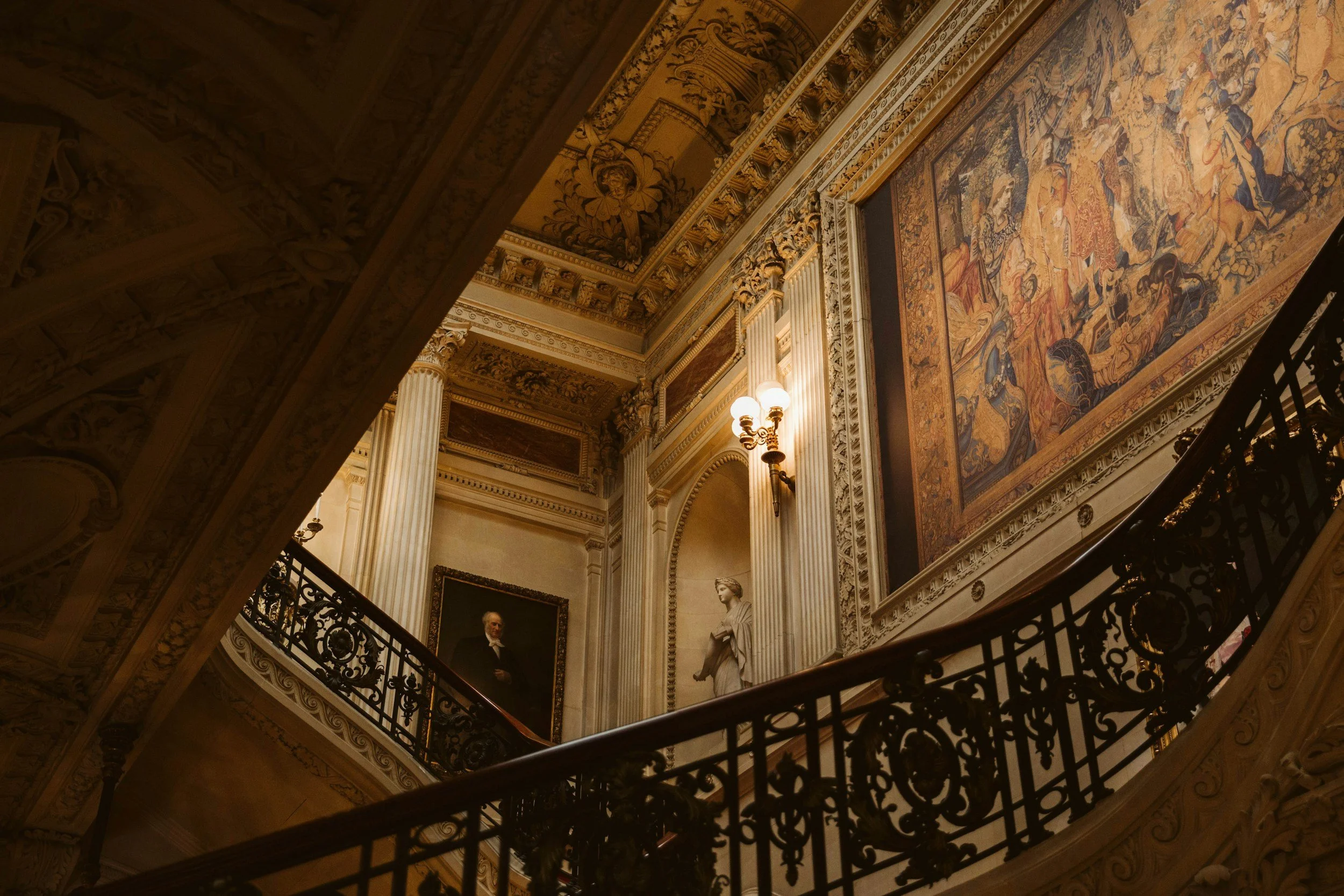Interior view of a grand, ornate building staircase with decorative railings, classical columns, and artwork on the walls and ceiling.