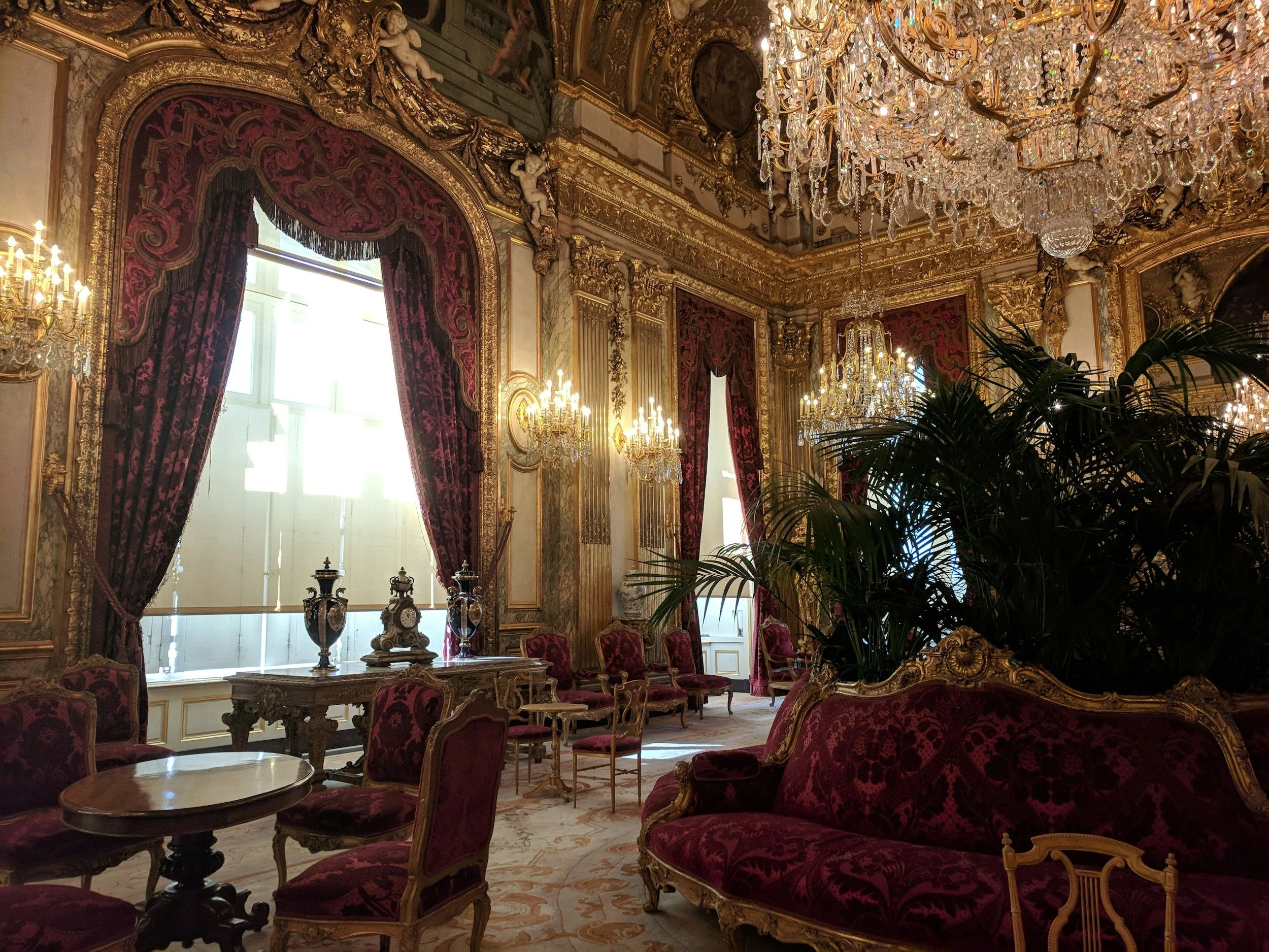 Luxurious 18th-century European style ballroom with ornate gold detailing, large chandeliers, red velvet curtains, and antique furniture.