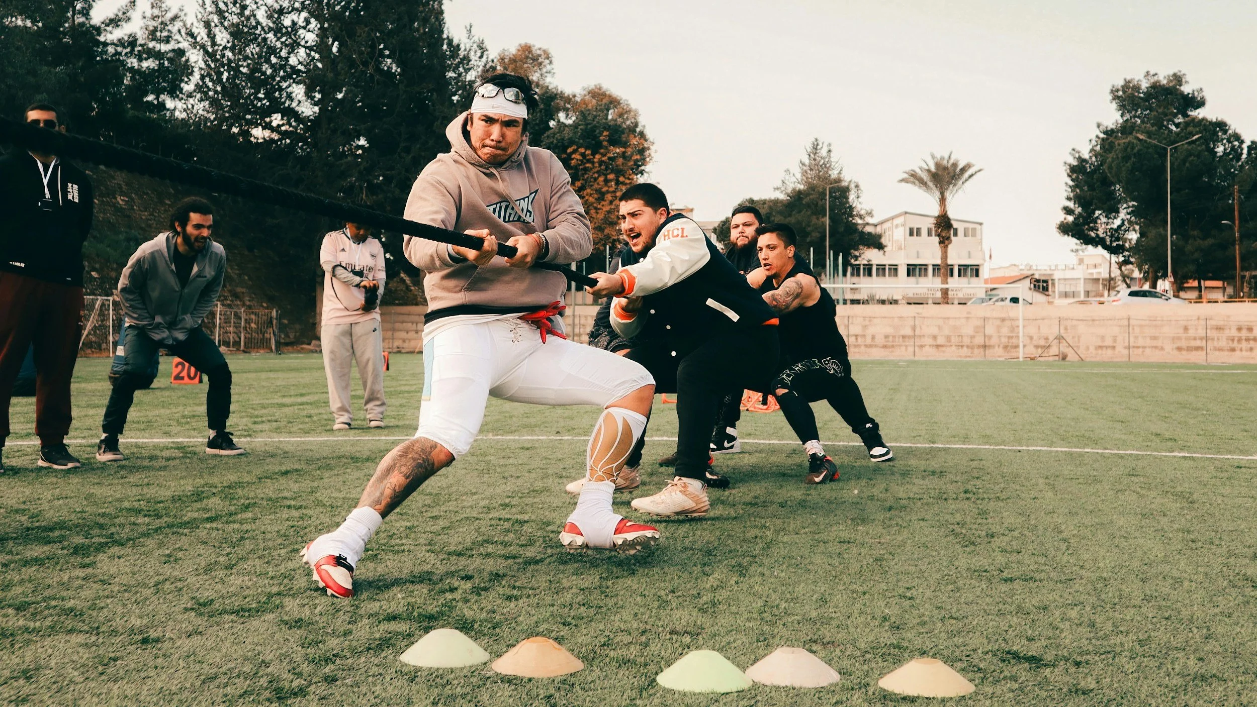 A group of men participate in a tug-of-war game on a sports field, with one man in the foreground pulling a rope, others cheering and watching.