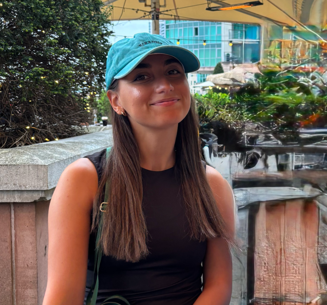 A woman wearing a light blue cap and a black sleeveless top, sitting outdoors with a smile, glass building and greenery in the background.