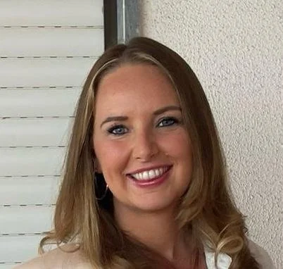 A smiling woman with blonde hair, wearing hoop earrings, standing indoors against a white wall and window blinds.