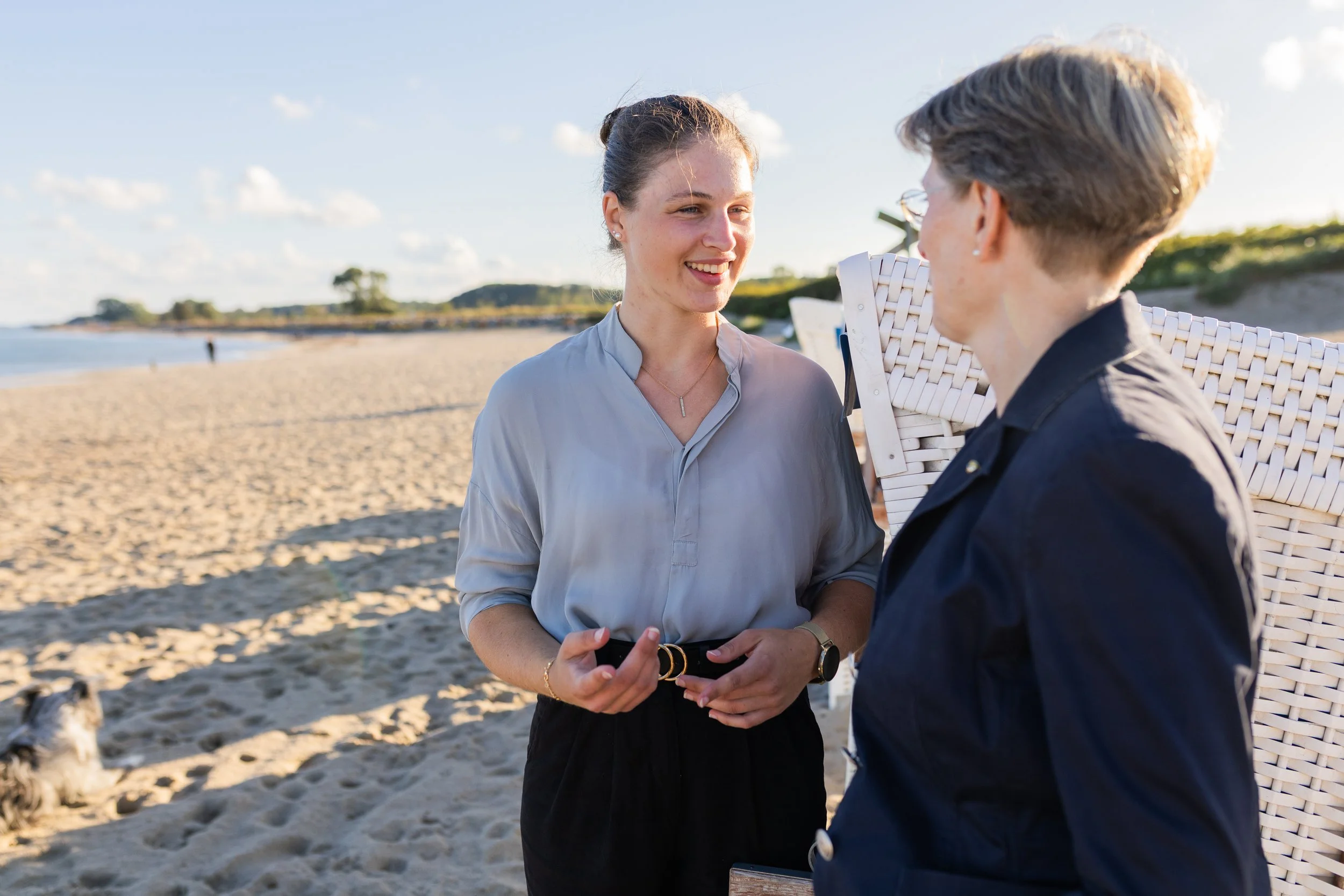 Two women talking on a beach during sunset, with sand, water, and distant trees in the background.