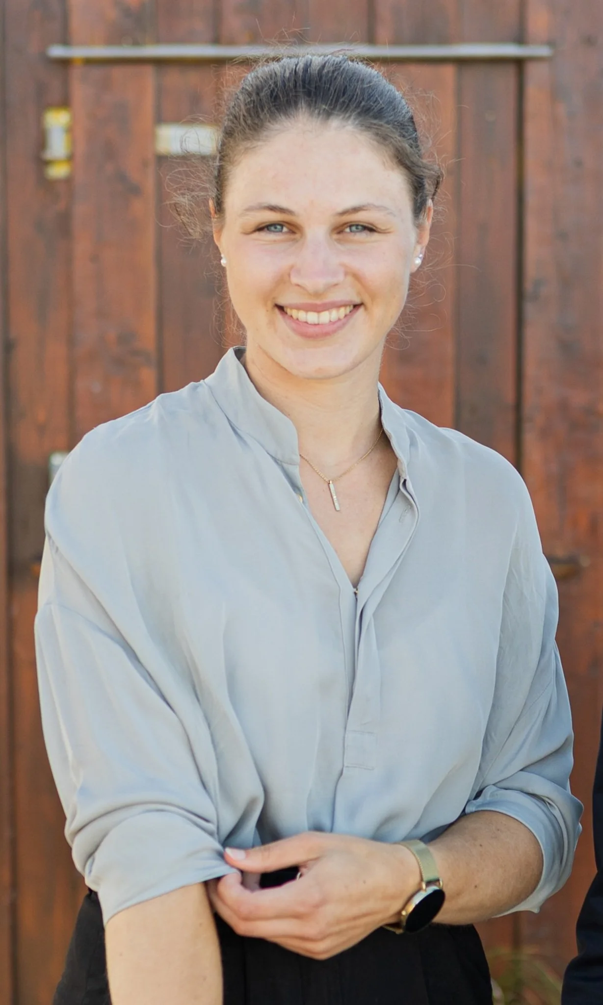 A woman with dark hair tied back, wearing a light-colored blouse, smiling, standing outdoors in front of a wooden fence.