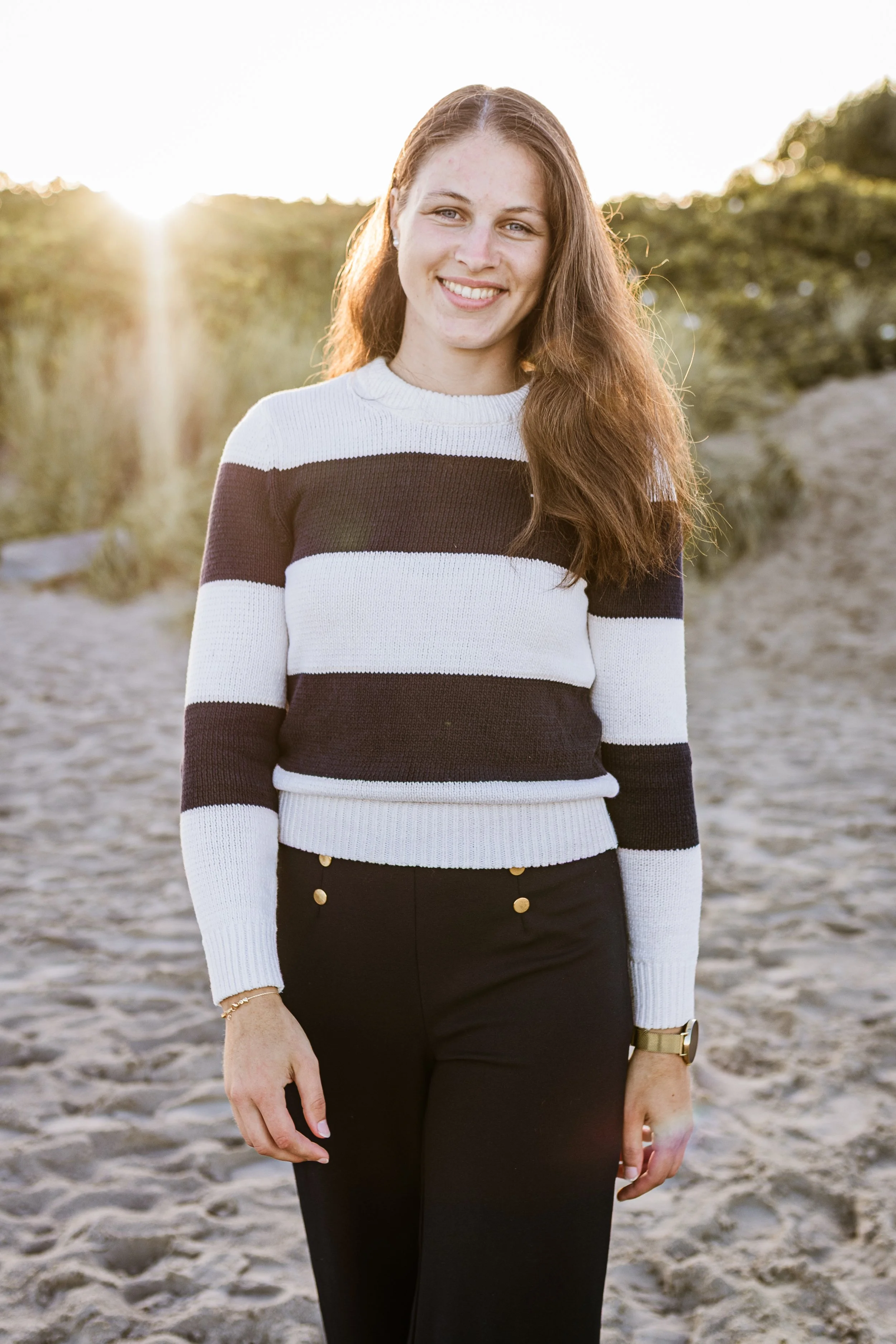 A smiling woman with long, wavy auburn hair, standing on a sandy beach at sunset, wearing a black and white striped sweater and black pants.