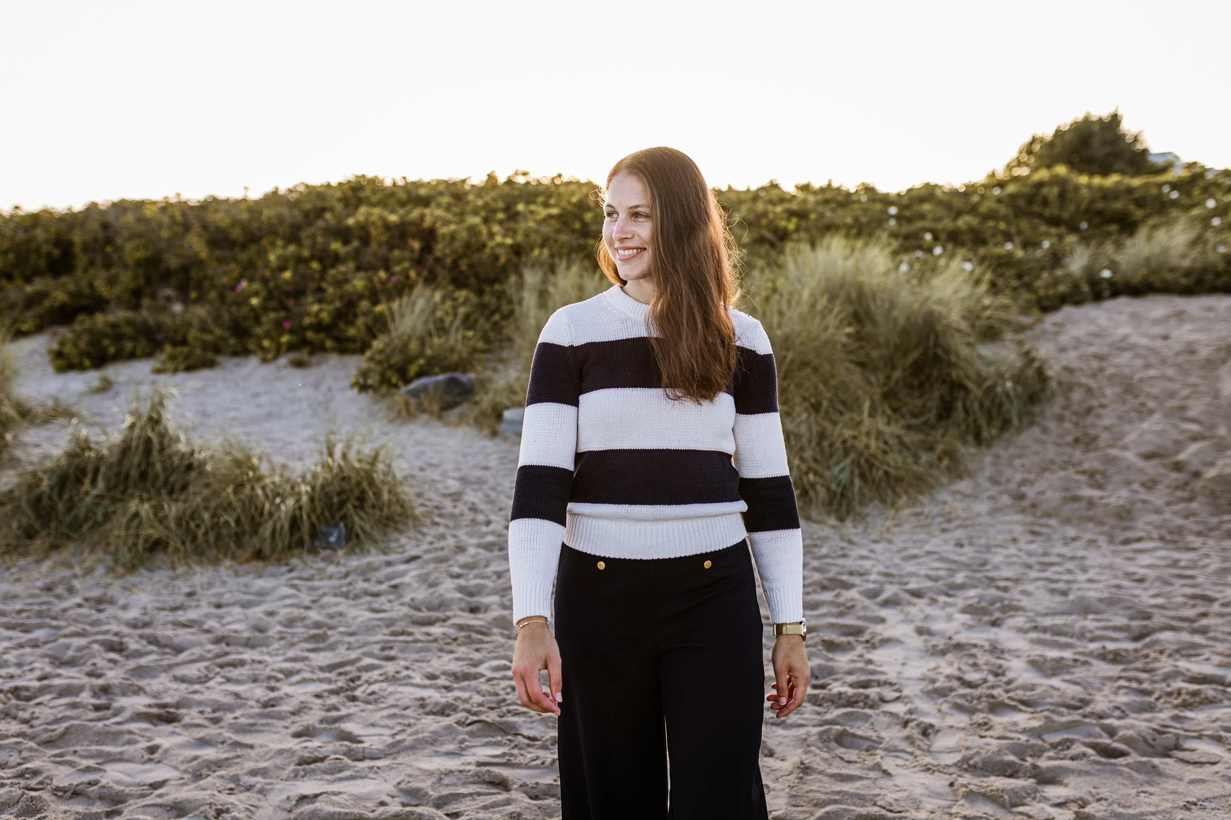 A smiling woman with long reddish-brown hair standing on a sandy beach with green shrubbery behind her, wearing a black and white striped sweater and black pants.