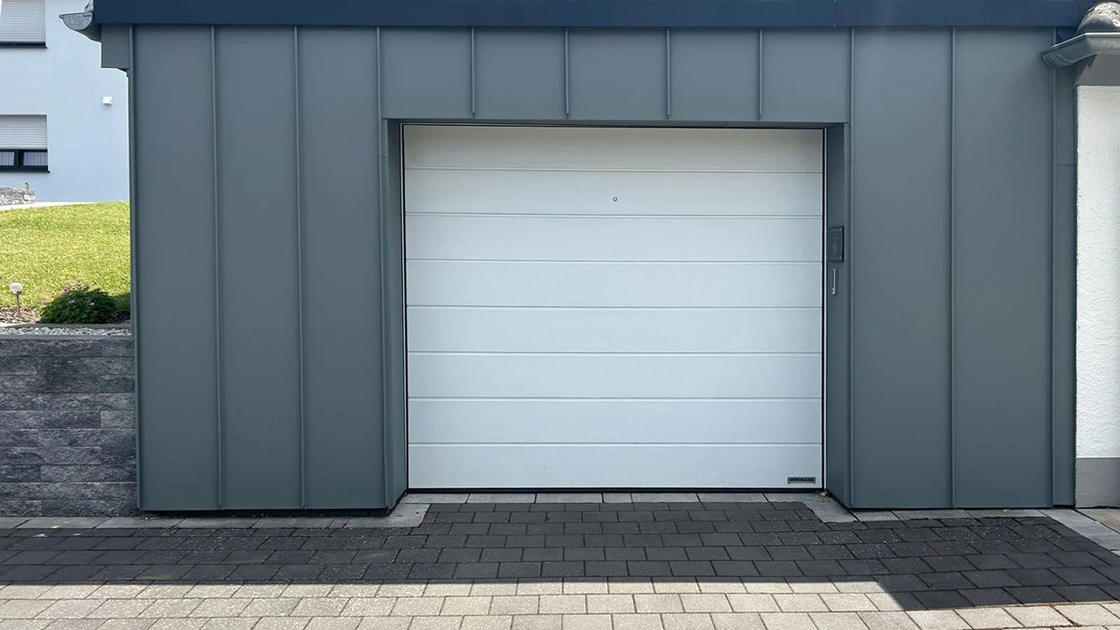 Ein graues Gebäude mit einer weißen Garagentür, daneben eine kleine Straßenfläche mit Pflastersteinen.