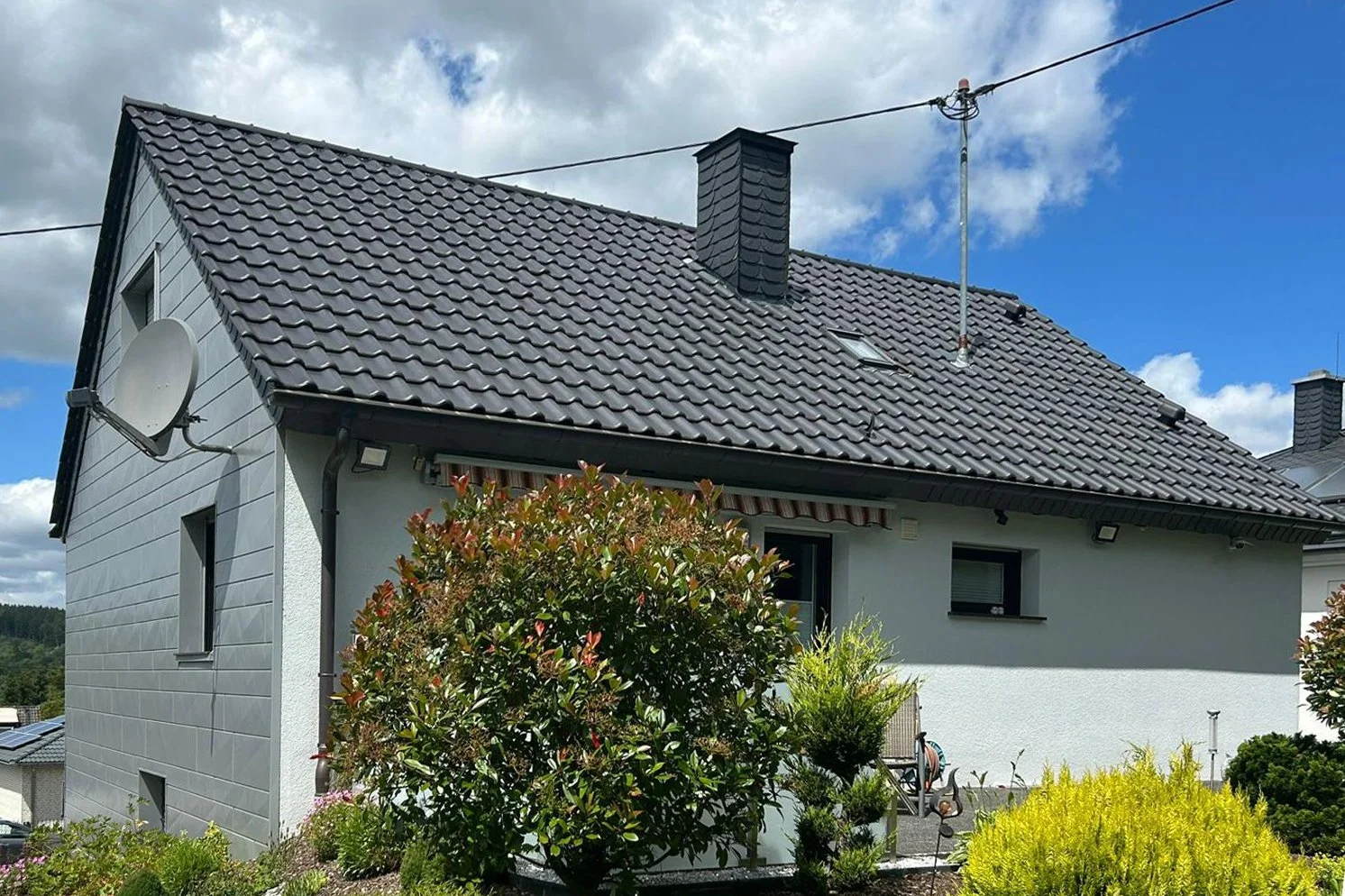 Ein Haus mit weißen Wänden und dunklen Dachziegeln, mit Satellitenschüssel und Antennen, umgeben von grünen Sträuchern und Pflanzen, bei sonnigem Himmel mit Wolken.