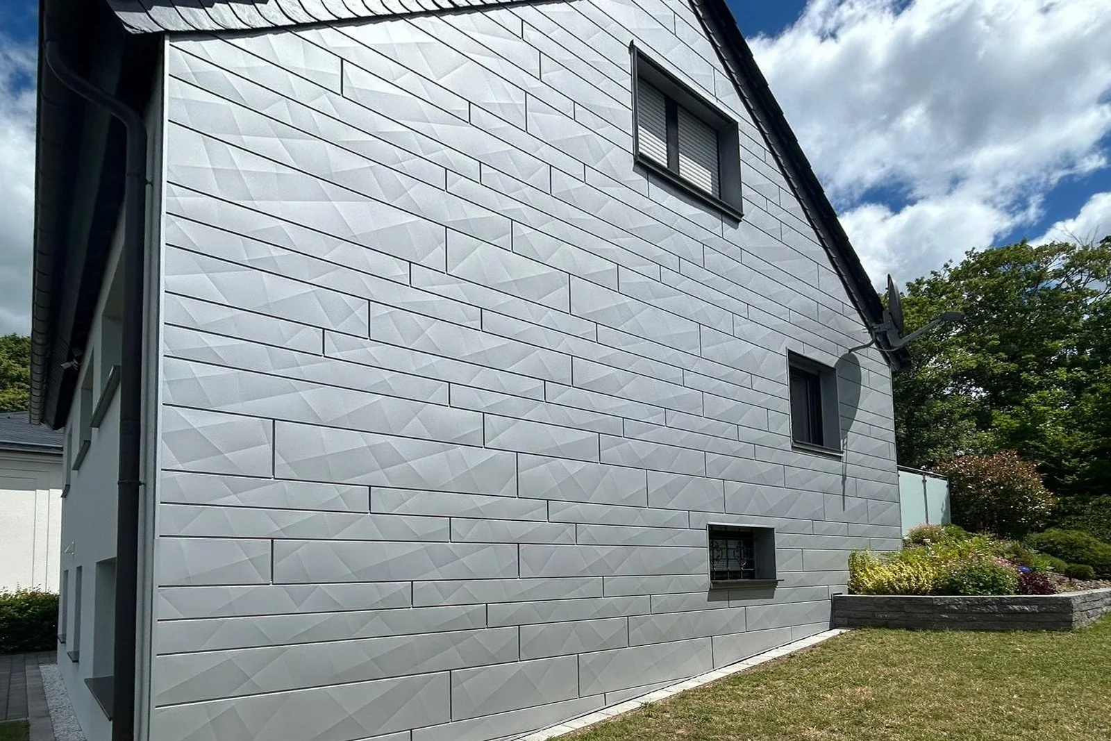 Das Bild zeigt die Außenfassade eines modernen Hauses mit mehreren kleinen Fenstern, die Verkleidung besteht aus dekorativen, geometrisch geformten weißen Paneelen. Es gibt einen Garten mit Blumen und Büschen daneben.