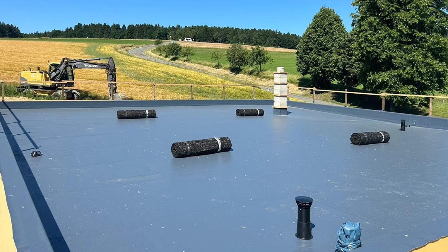 Dach mit Asphaltabdichtung, auf dem Rollrinnen und Lüftungsrohre installiert sind, mit grüner Landschaft und Bäumen im Hintergrund