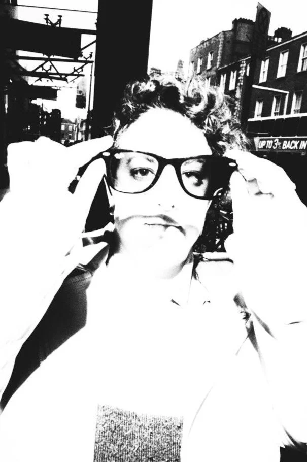 Person adjusting glasses outdoors in a city, with buildings and a sign in the background, in black and white.