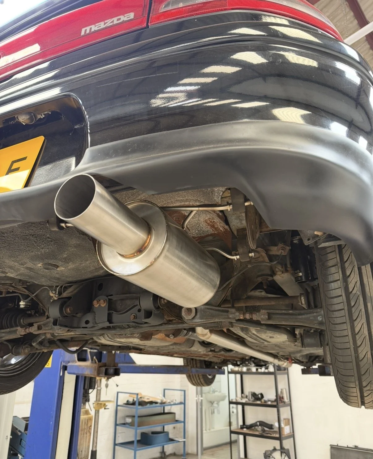 View of the underside of a black Mazda car with a silver exhaust pipe in an automotive repair shop, with shelves and equipment visible in the background.