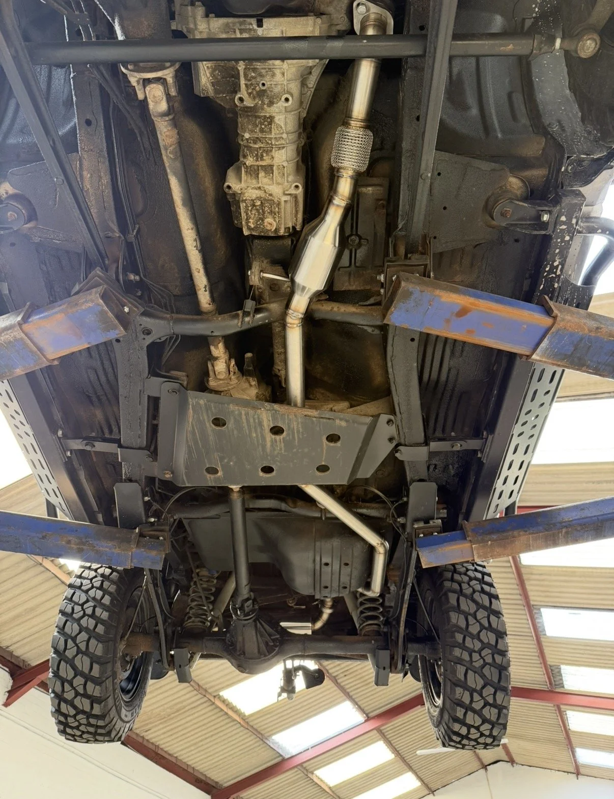 Underneath view of a lifted off-road vehicle showing the chassis, suspension, and exhaust system with the vehicle supported by metal beams.