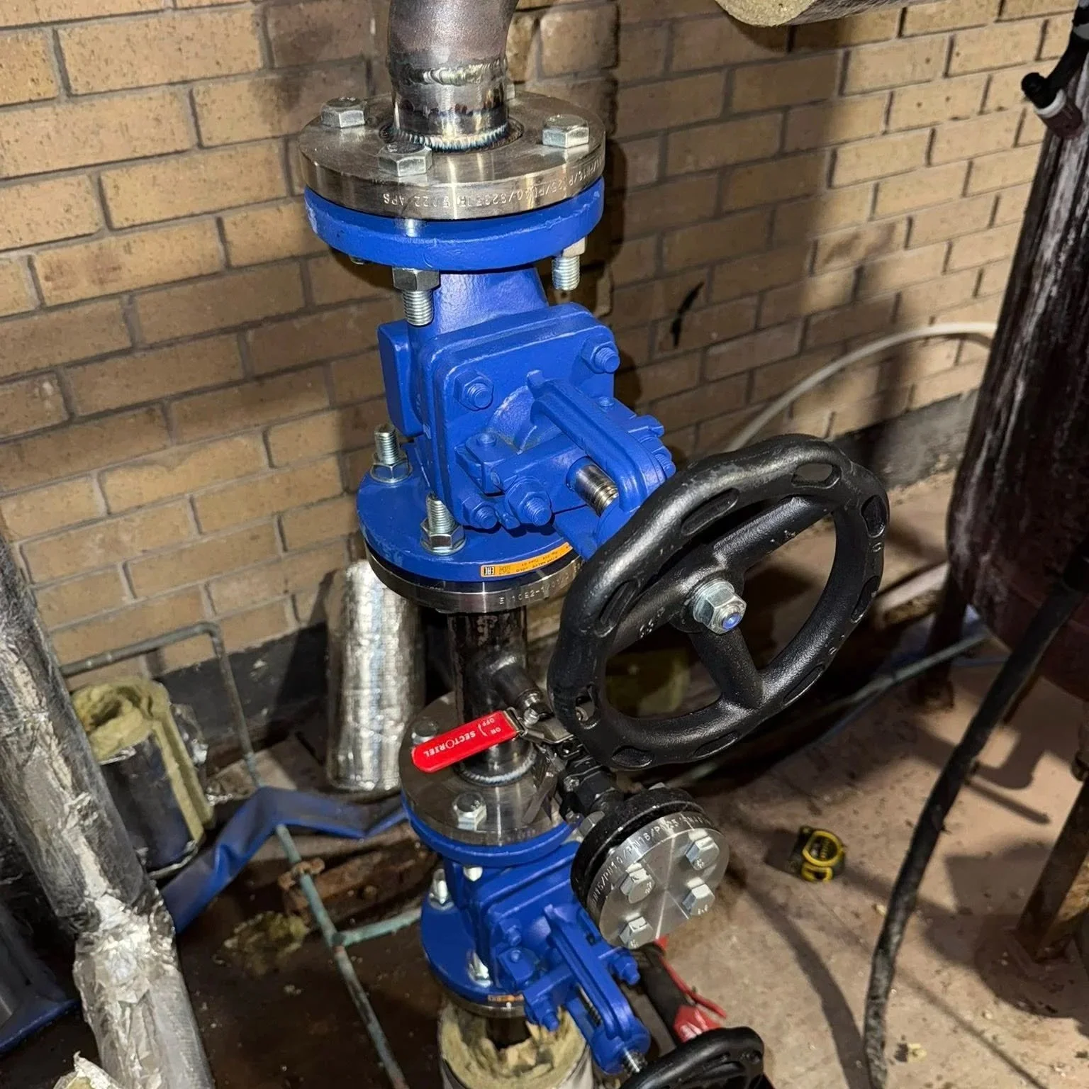 Close-up of a plumbing system with a blue valve, black wheel handle, and metallic pipes against a brick wall.