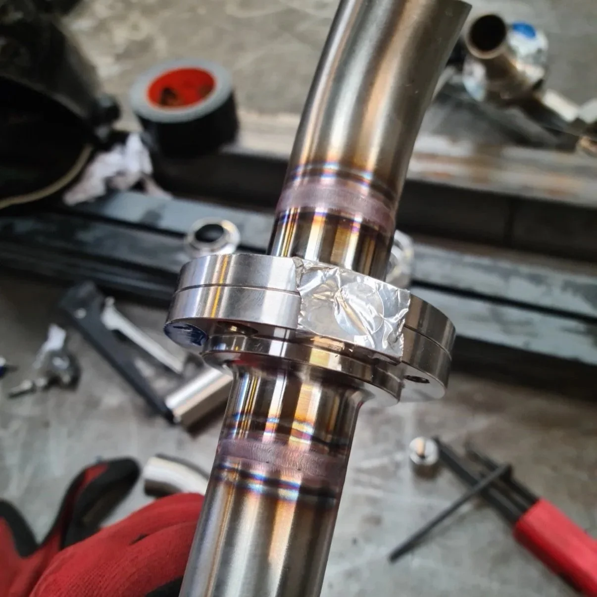 Close-up of a metal shaft with a ring and tape applied, likely part of a mechanical or automotive assembly, with tools and parts in the background.
