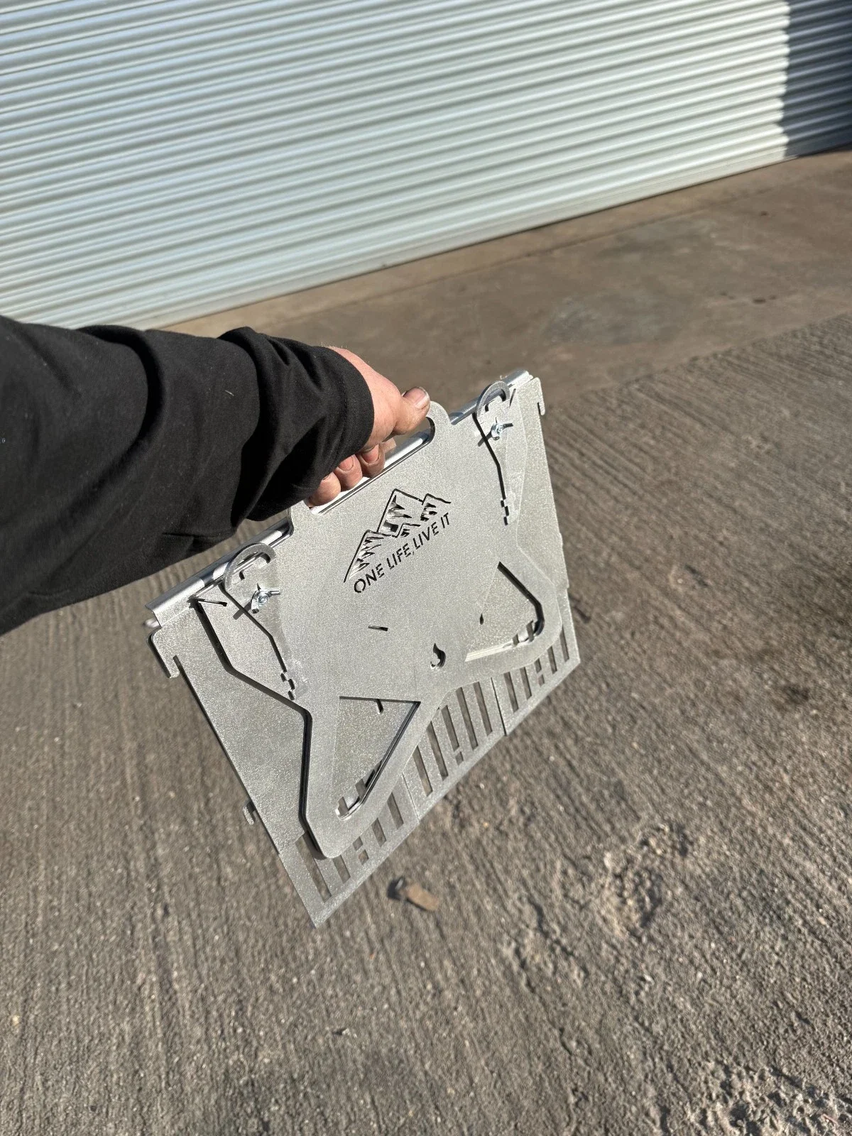 Person holding a metal camping stove or grill with the phrase "ONE LIFE LIVE IT" and mountain logo, in front of a closed metal roll-up door on a concrete surface.