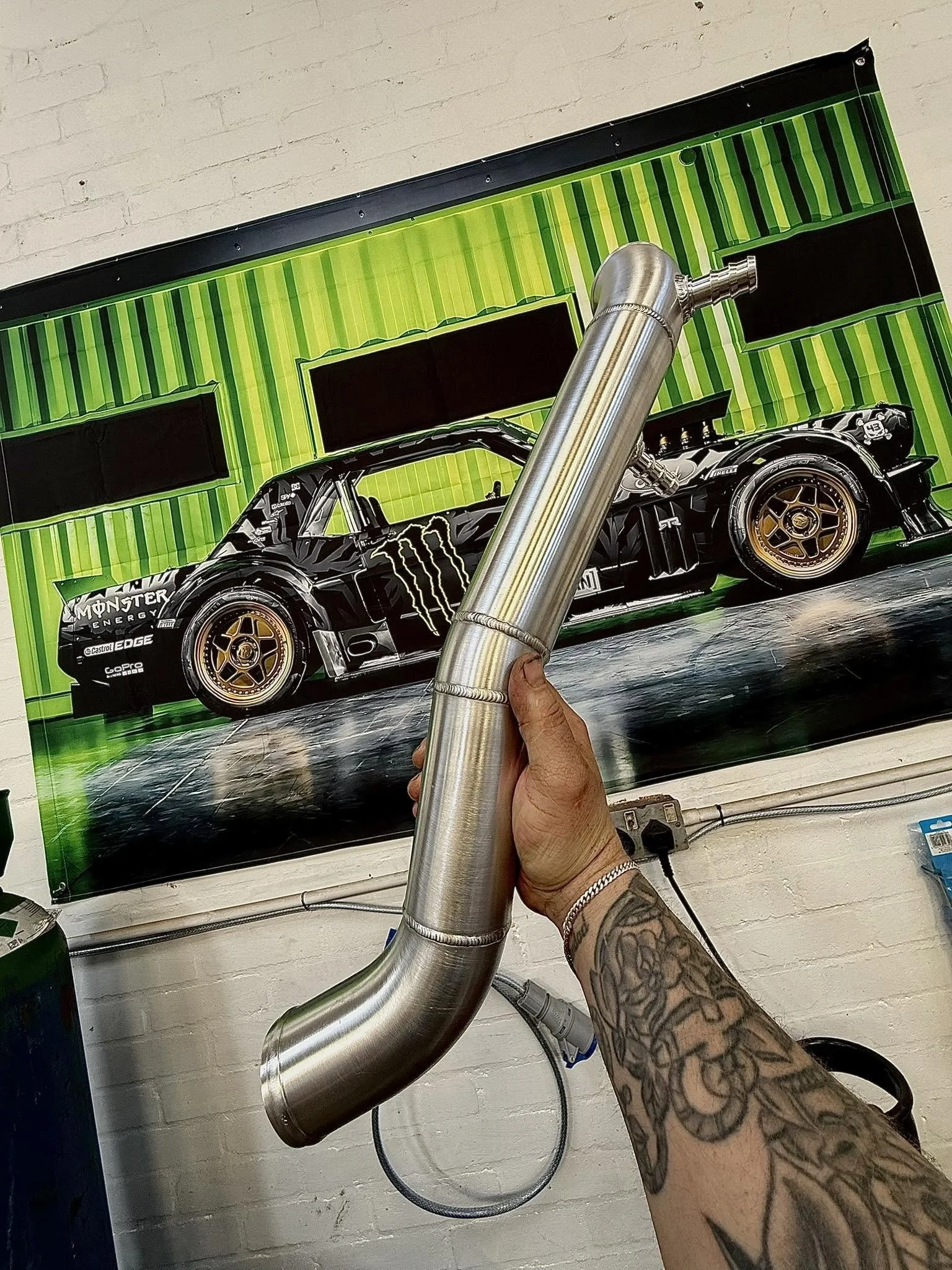 A tattooed arm holding a large metallic pipe in front of a colorful poster featuring a racing car with Monster Energy branding and a bright green, yellow, and black striped background.