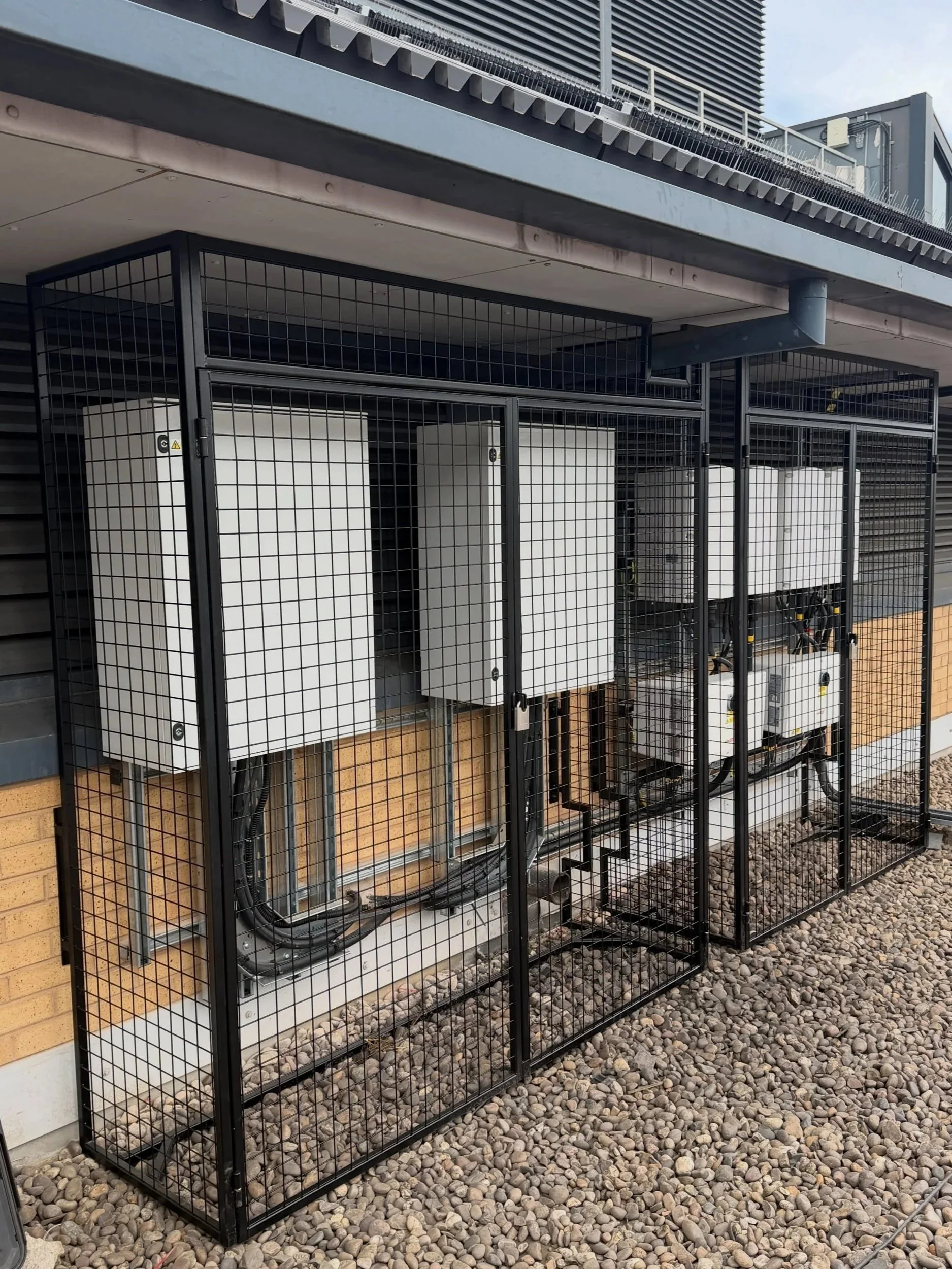 Outdoor utility area with electrical equipment enclosed in black metal fencing, set against a building with a brick wall and gravel ground.
