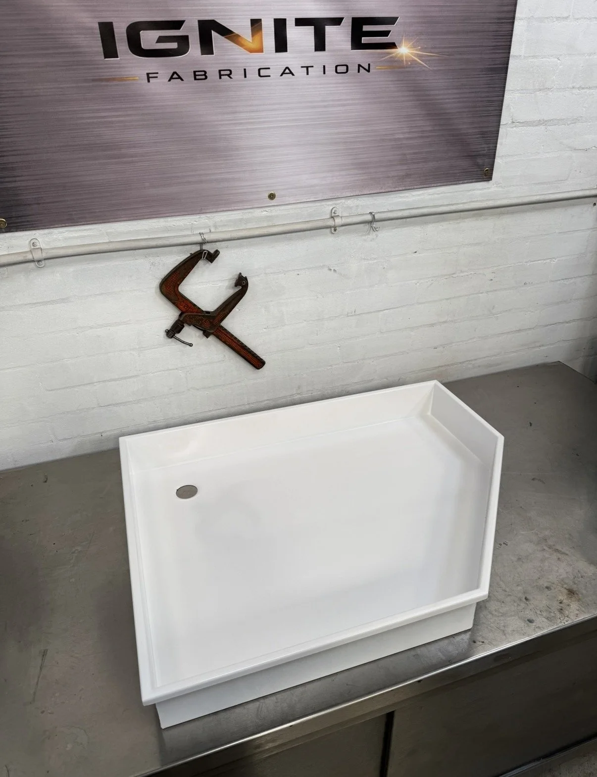 A white shower tray sitting on a metal surface in a manufacturing or assembly area, with a wall-mounted clamp tool hanging above and a sign reading "IGNITE FABRICATION" on the wall.