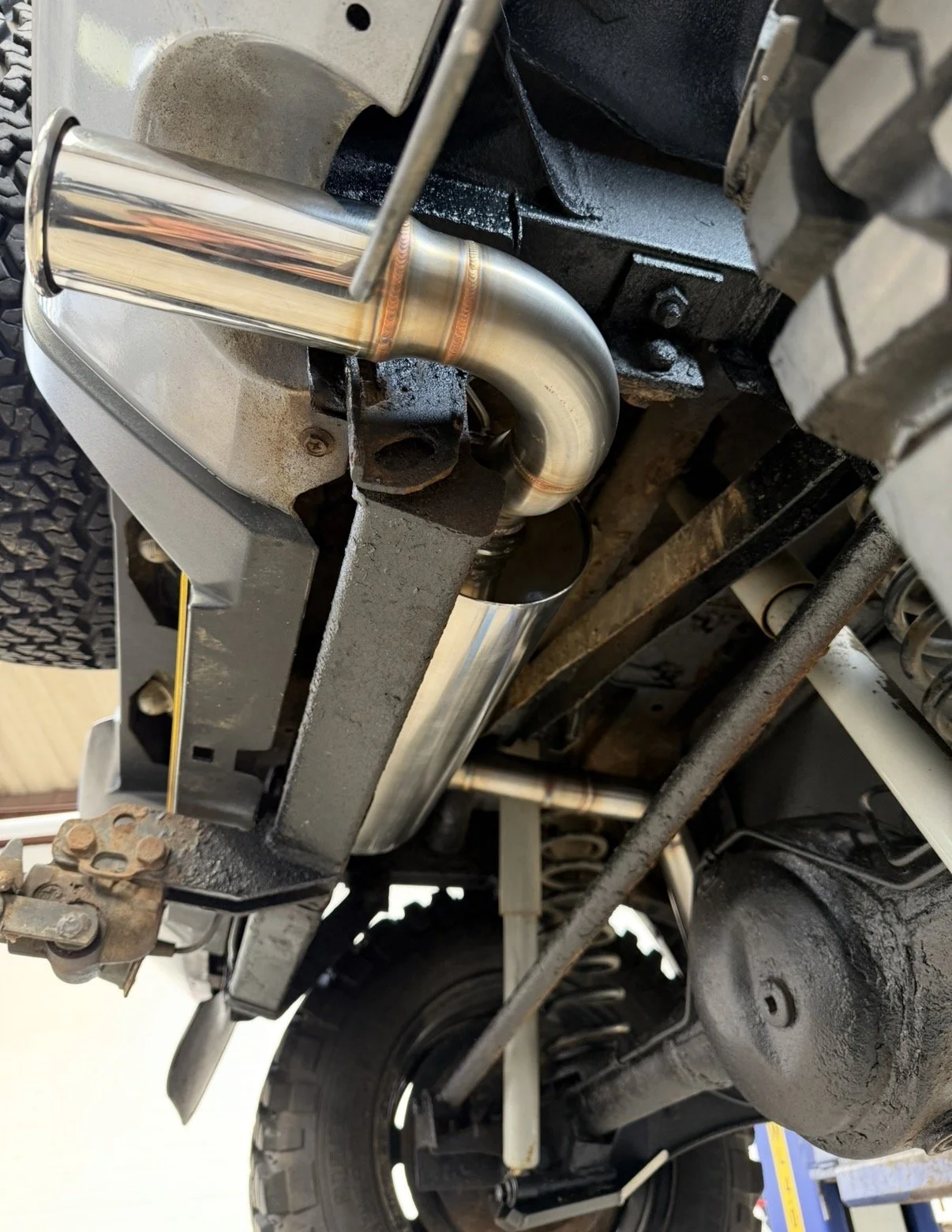 Underneath view of a vehicle's exhaust system showing a shiny new metal exhaust pipe and muffler, surrounded by various mechanical parts and a rusty frame.