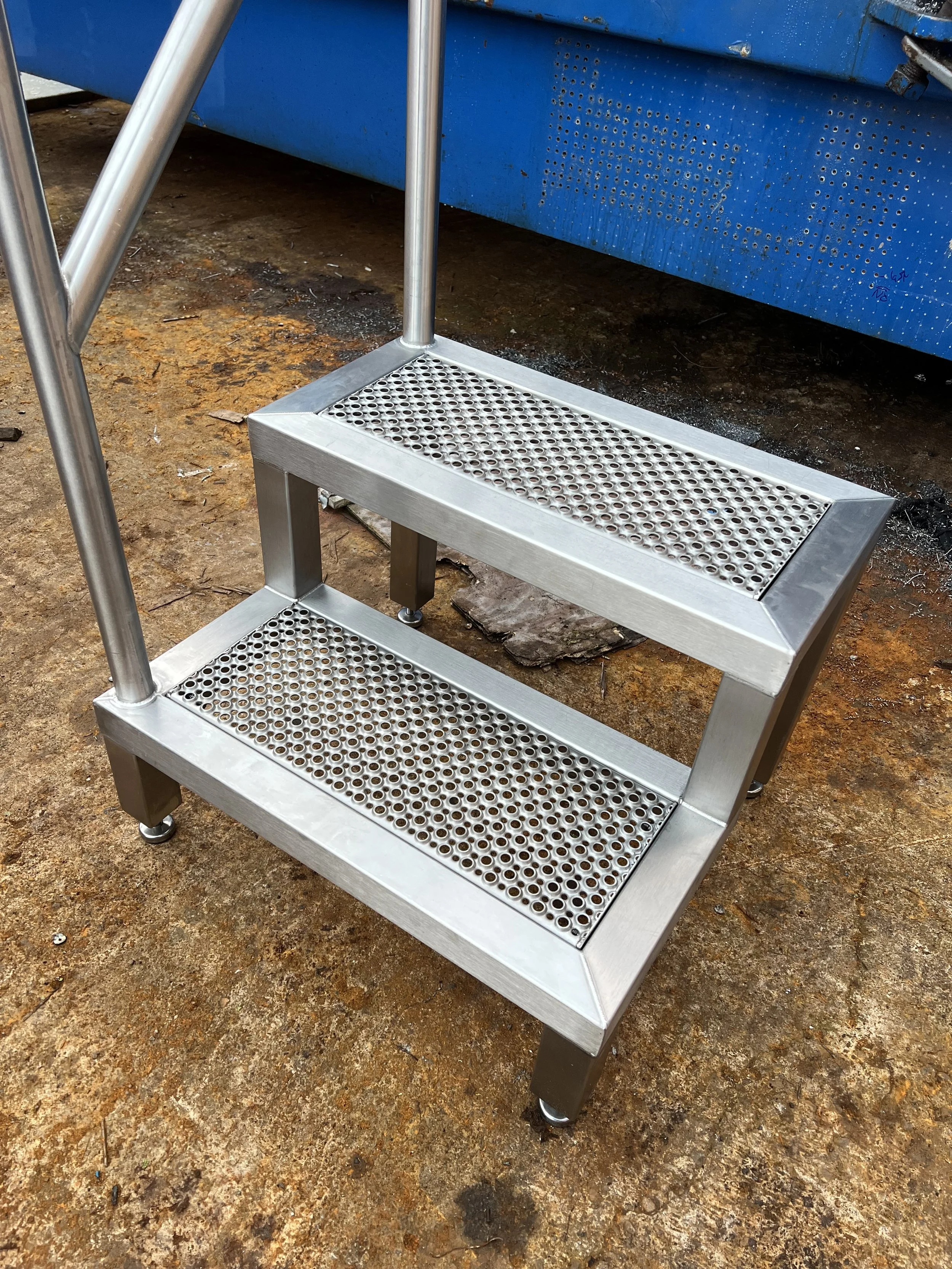 A small, two-tiered metal rolling cart with perforated shelves and black rubber feet, placed outdoors on a dirt and rock surface near a blue structure.
