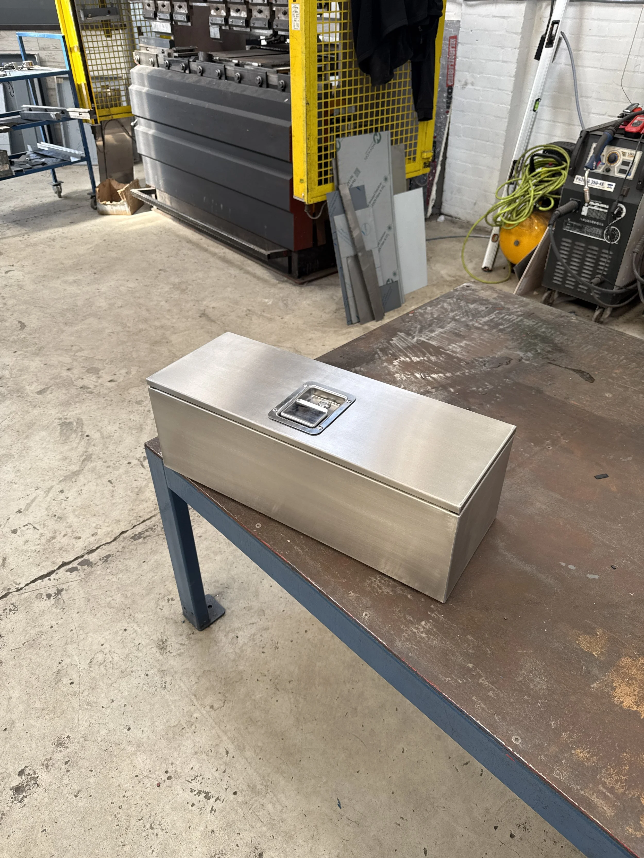 A metal box with a lid on a workbench in an industrial workshop. In the background, there are metal shelves, a welding machine with hoses, a yellow safety guard around machinery, and various tools and materials.