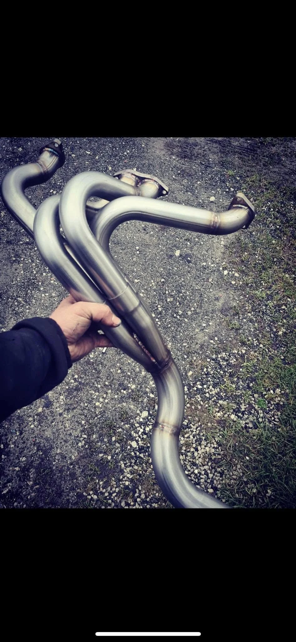 A person holds a shiny, metallic, custom exhaust pipe for a vehicle, with a gravel and dirt ground in the background.