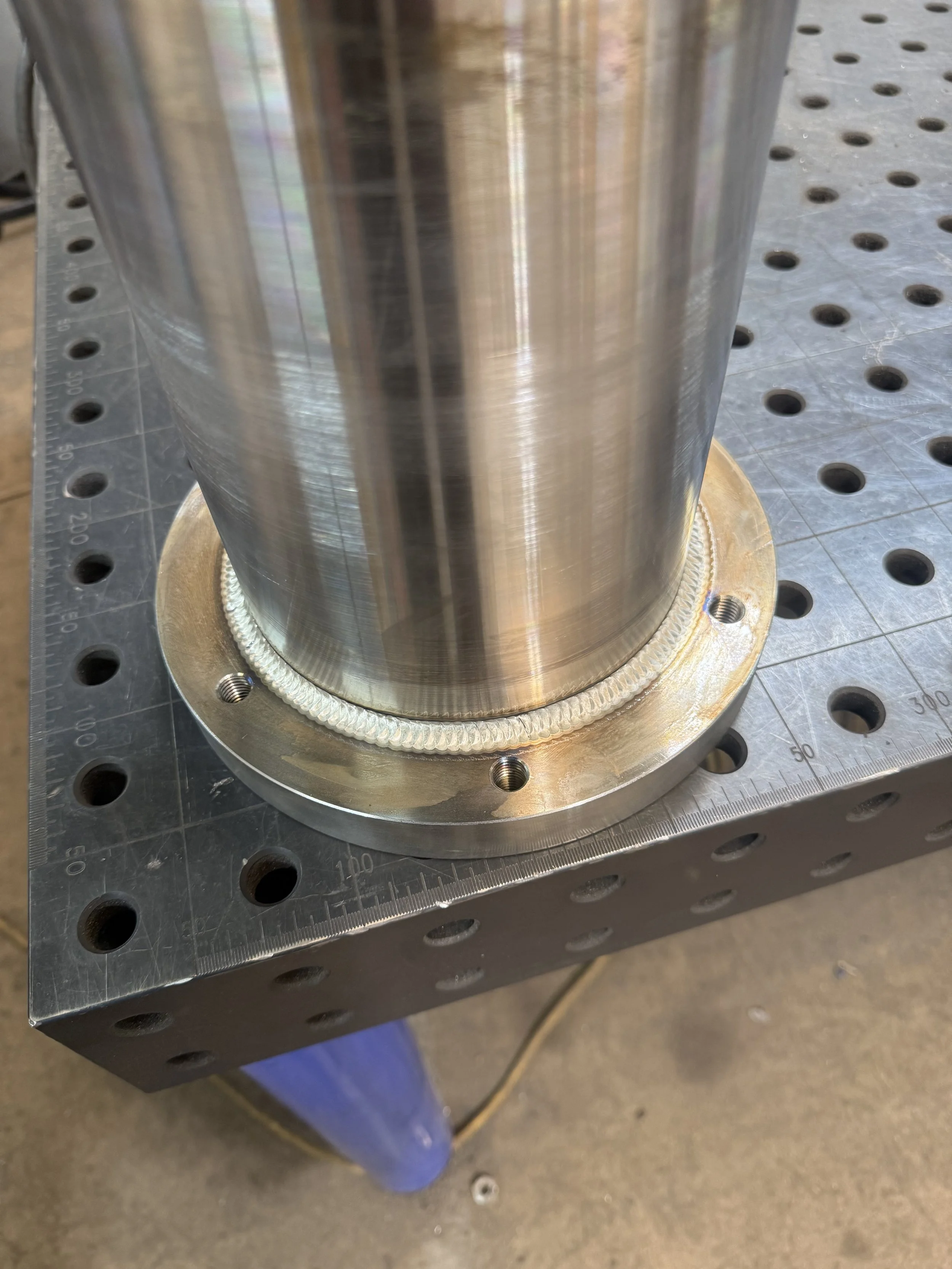 Close-up of a large, shiny metal cylindrical object mounted on a table with measurement markings, possibly an industrial or mechanical component.