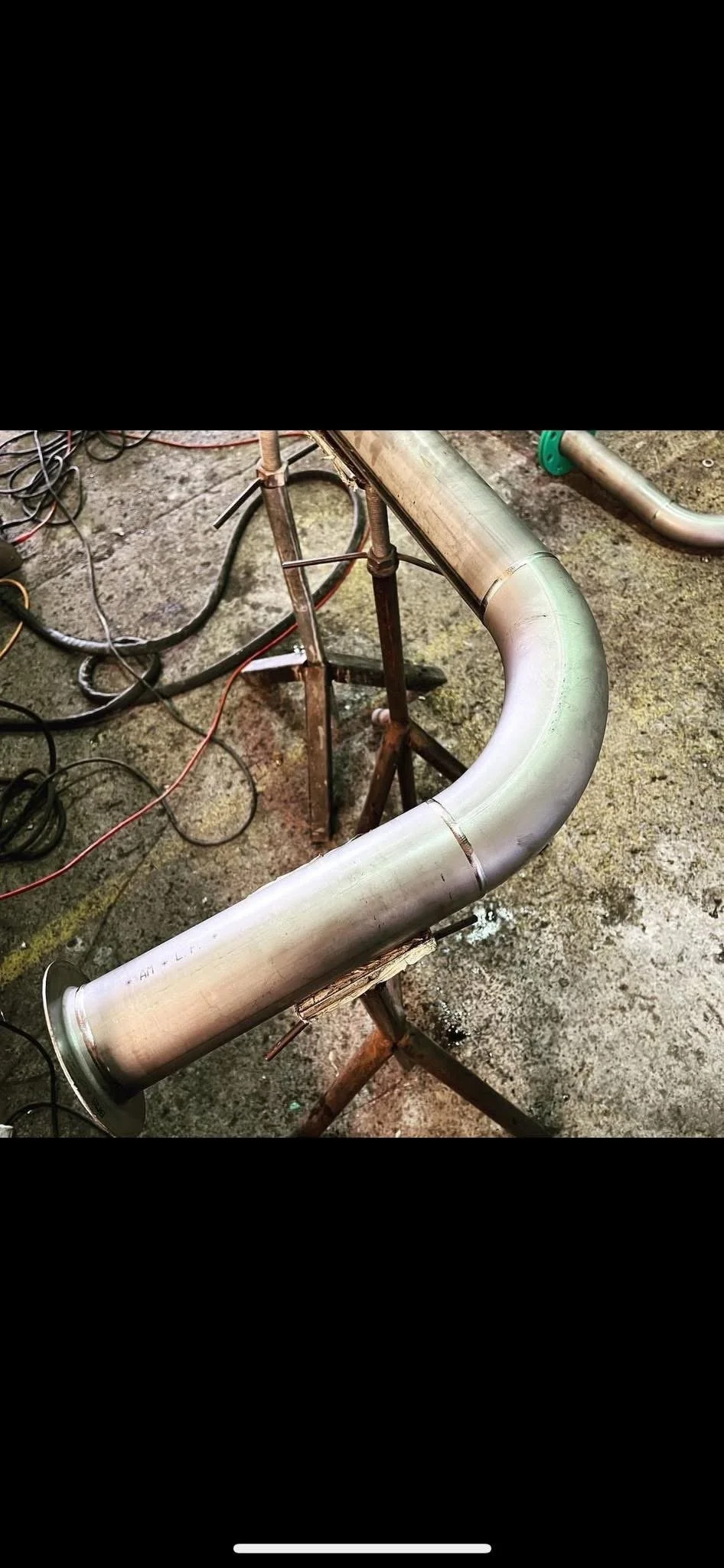 A metal pipe with a bend resting on a rusty metal stand on a concrete floor, surrounded by wires and tools.