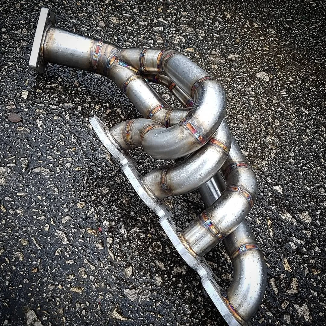 A metal exhaust manifold with multiple curved pipes lies on a wet asphalt surface.
