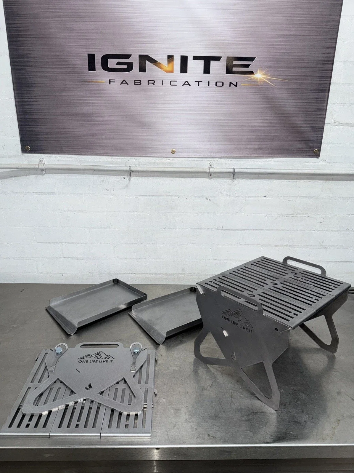Metal grilling equipment and trays on a stainless steel countertop with a sign that reads 'IGNITE FABRICATION' on a white brick wall in the background.